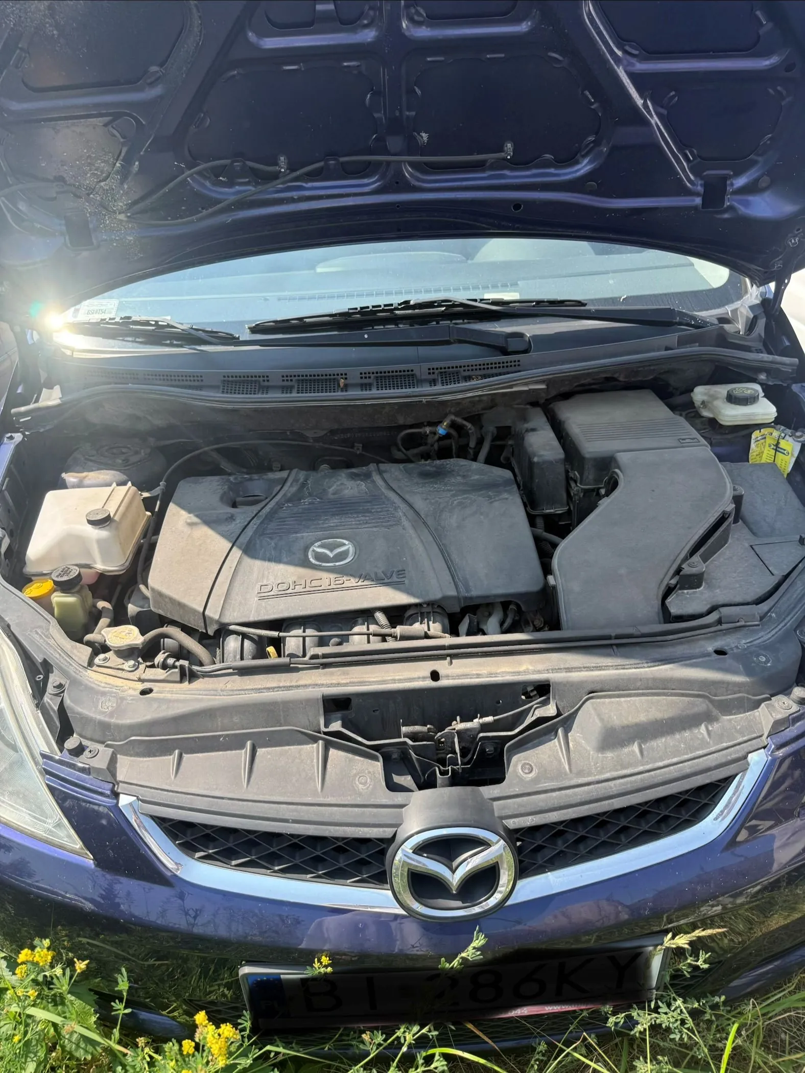 Widok z przodu na otwartą maskę ciemnoniebieskiego samochodu marki Mazda. Widoczny jest silnik z plastikową osłoną z logo Mazdy i napisem 'DOHC 16-VALVE'. Poniżej znajduje się grill z dużym logo Mazdy oraz fragment tablicy rejestracyjnej z tekstem 'BI 286KY'.