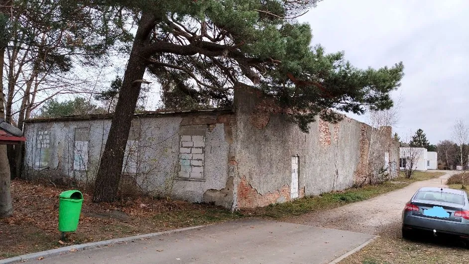 Zdjęcie przedstawiające zniszczony, jednopiętrowy budynek z szarego betonu i cegieł z odpadającym tynkiem. Budynek ma kilka okien, z których niektóre są zamurowane białymi cegłami. Przed budynkiem rośnie duże drzewo iglaste, którego gałęzie zwisają nad dachem. Po lewej stronie znajduje się zielony kosz na śmieci, a po prawej zaparkowany jest srebrny samochód osobowy na piaszczystej drodze.