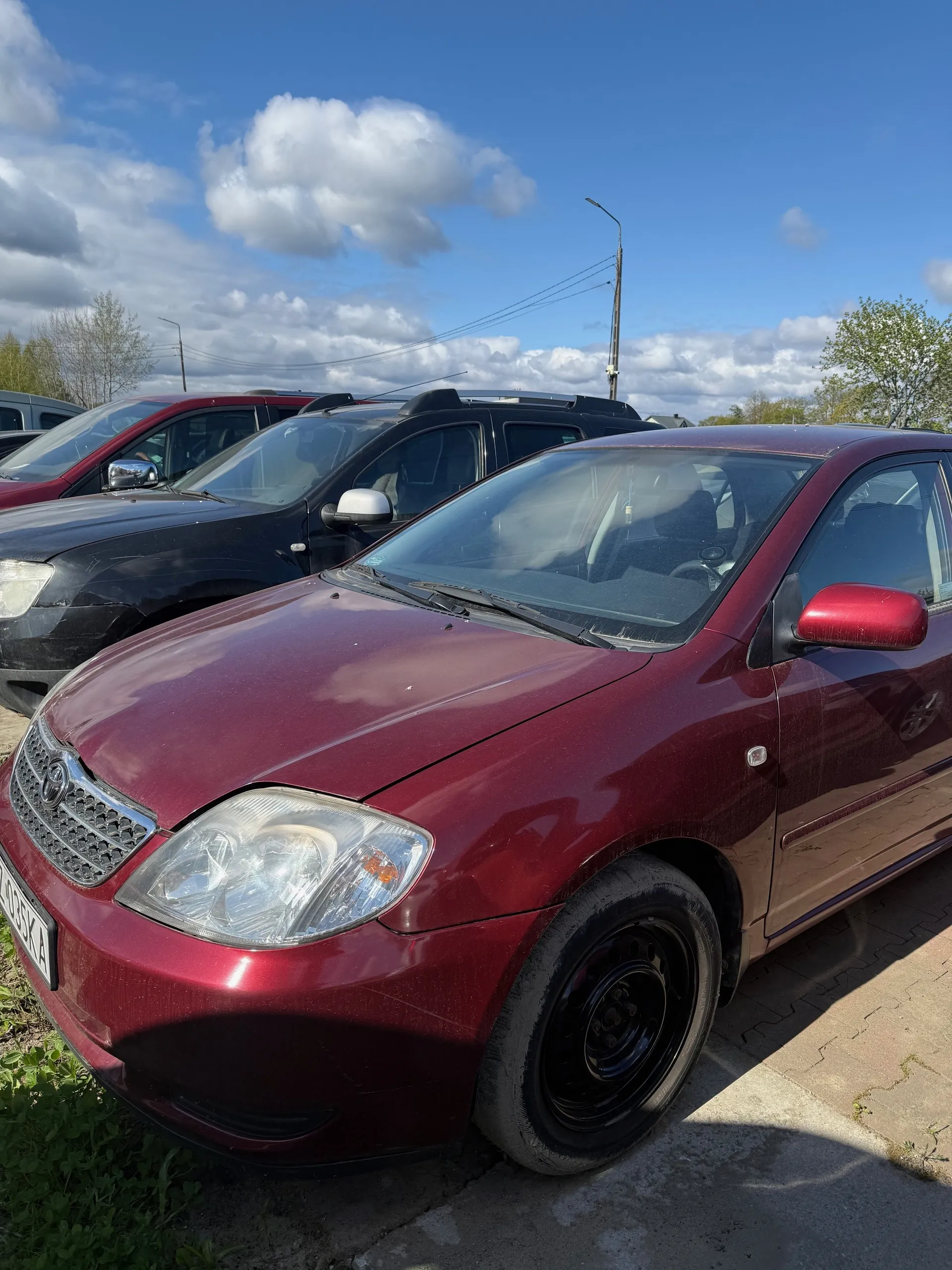 Czerwony samochód marki Toyota na pierwszym planie, z tablicą rejestracyjną 493KA. Za nim widoczne są czarny SUV i czerwony samochód. Tło stanowi niebo z białymi chmurami, drzewa oraz słup z przewodami elektrycznymi. Samochody zaparkowane są na nawierzchni z płytek i trawie.