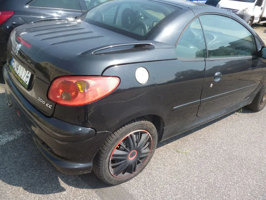 Czarny samochód Peugeot 206 CC zaparkowany na asfaltowym placu. Widoczny jest tył i prawy bok pojazdu. Na klapie bagażnika znajduje się napis '206 CC' oraz tablica rejestracyjna. Samochód ma czarne felgi z czerwonym obramowaniem.