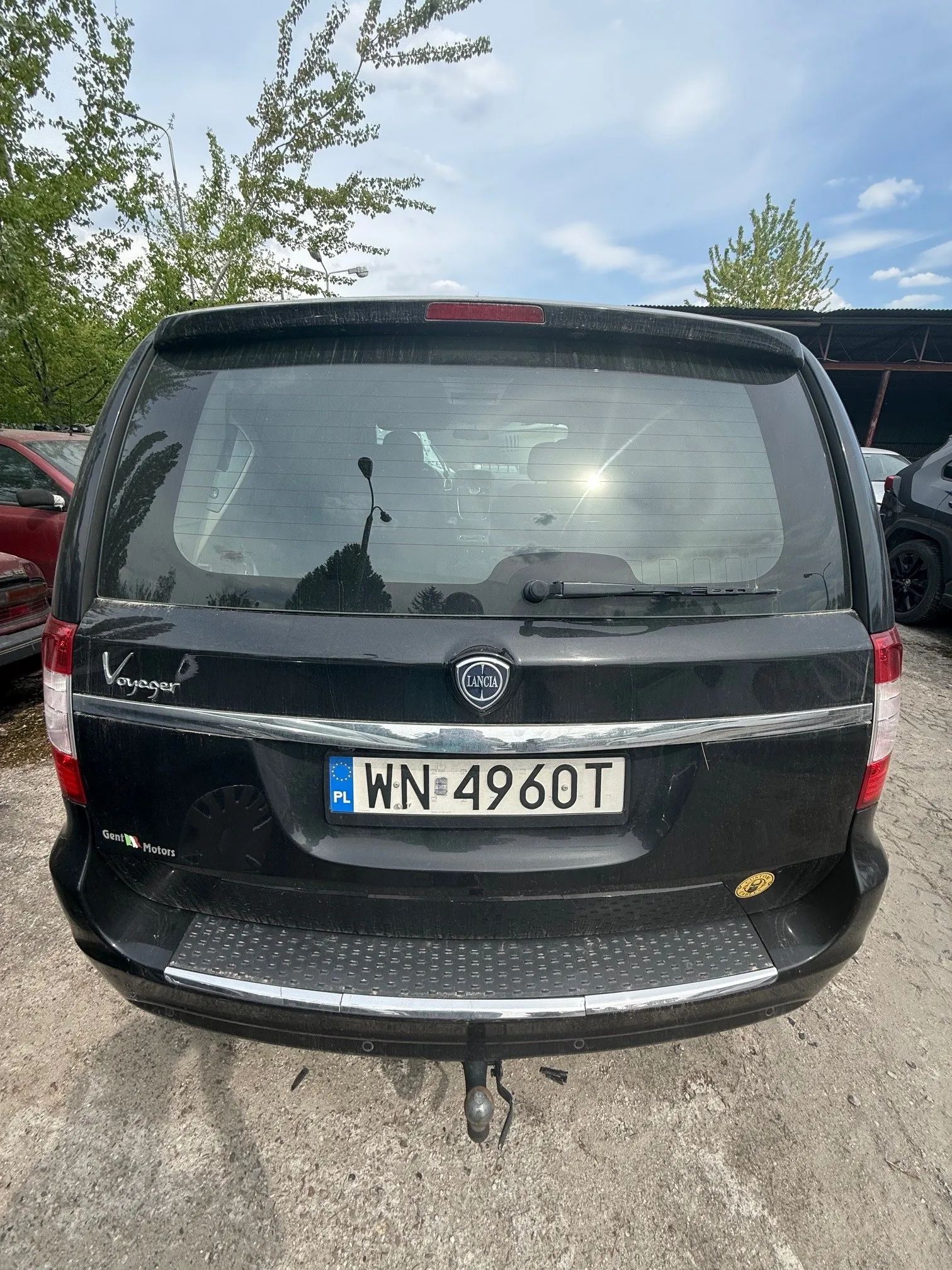 Czarny samochód osobowy marki Lancia Voyager widziany od tyłu. Na bagażniku widoczne napisy: "Voyager" po lewej stronie, logo "Lancia" w centrum oraz tablica rejestracyjna "PL WN 4960T" z niebieskim pasem po lewej stronie. Poniżej tablicy znajduje się napis "Gent Motors". W tle drzewa, częściowo widoczne inne pojazdy oraz niebo z chmurami. Samochód stoi na nierównym, żwirowym podłożu.