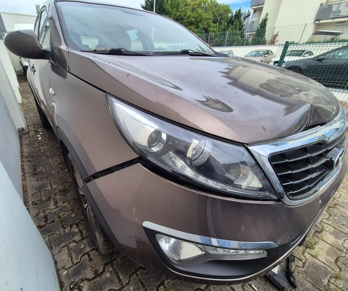 Zdjęcie przodu brązowego samochodu typu SUV, zaparkowanego na kostce brukowej. Pojazd jest uszkodzony – widoczne jest wgniecenie na masce nad grillem oraz szczelina między zderzakiem a błotnikiem po prawej stronie obrazu. W tle widoczne są inne samochody, zielone drzewa oraz fragmenty białych budynków mieszkalnych.
