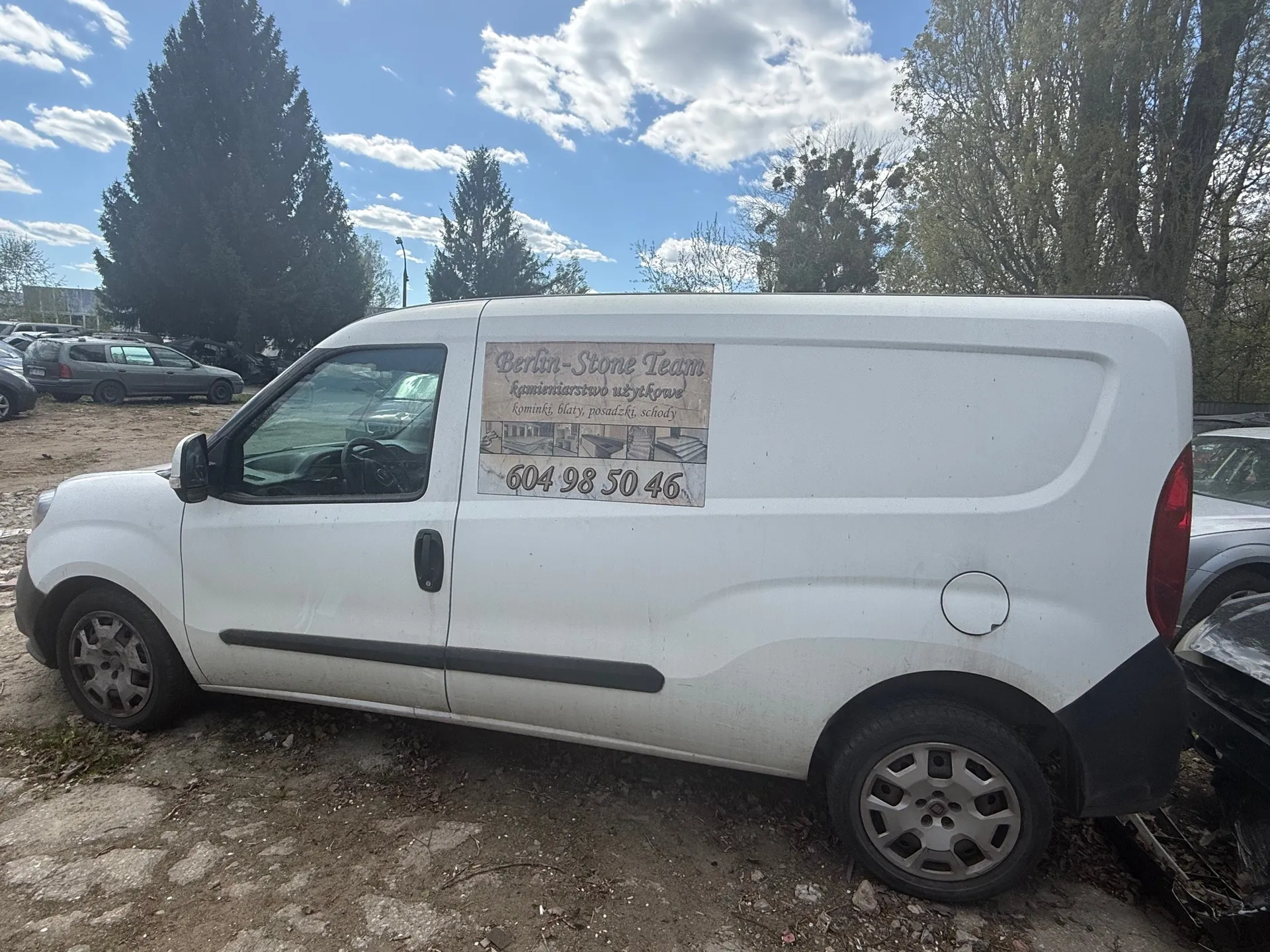 Biały furgon zaparkowany na brudnej powierzchni z innymi pojazdami na tle. Na boku furgonu znajduje się tablica z napisem: "Berlin-Stone Team, kamieniarstwo użytkowe, kominki, blaty, posadzki, schody, 604 98 50 46". W tle widoczne są drzewa, niebo z rozproszonymi chmurami oraz dodatkowe zaparkowane samochody. Furgon skierowany jest w lewo, z widocznymi przednim i tylnym kołem.