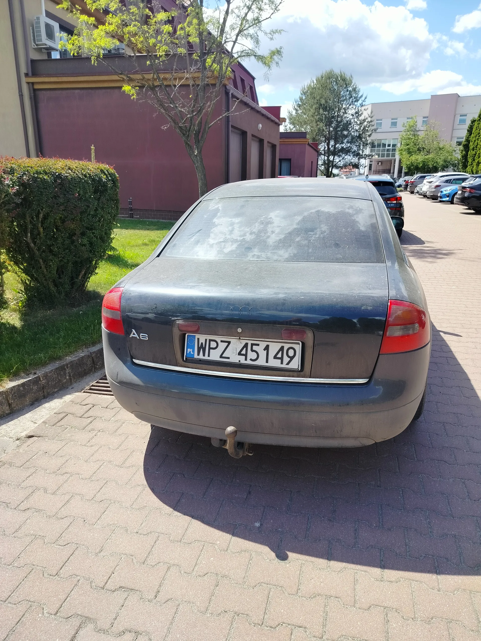 Tylne widok ciemnoszarego samochodu Audi A6 zaparkowanego na brukowanym parkingu. Widoczna tablica rejestracyjna PL WPZ 45149. Samochód jest nieco zabrudzony. W tle budynki, drzewa, krzewy i częściowo zachmurzone niebo. Po prawej stronie kolejne zaparkowane pojazdy.