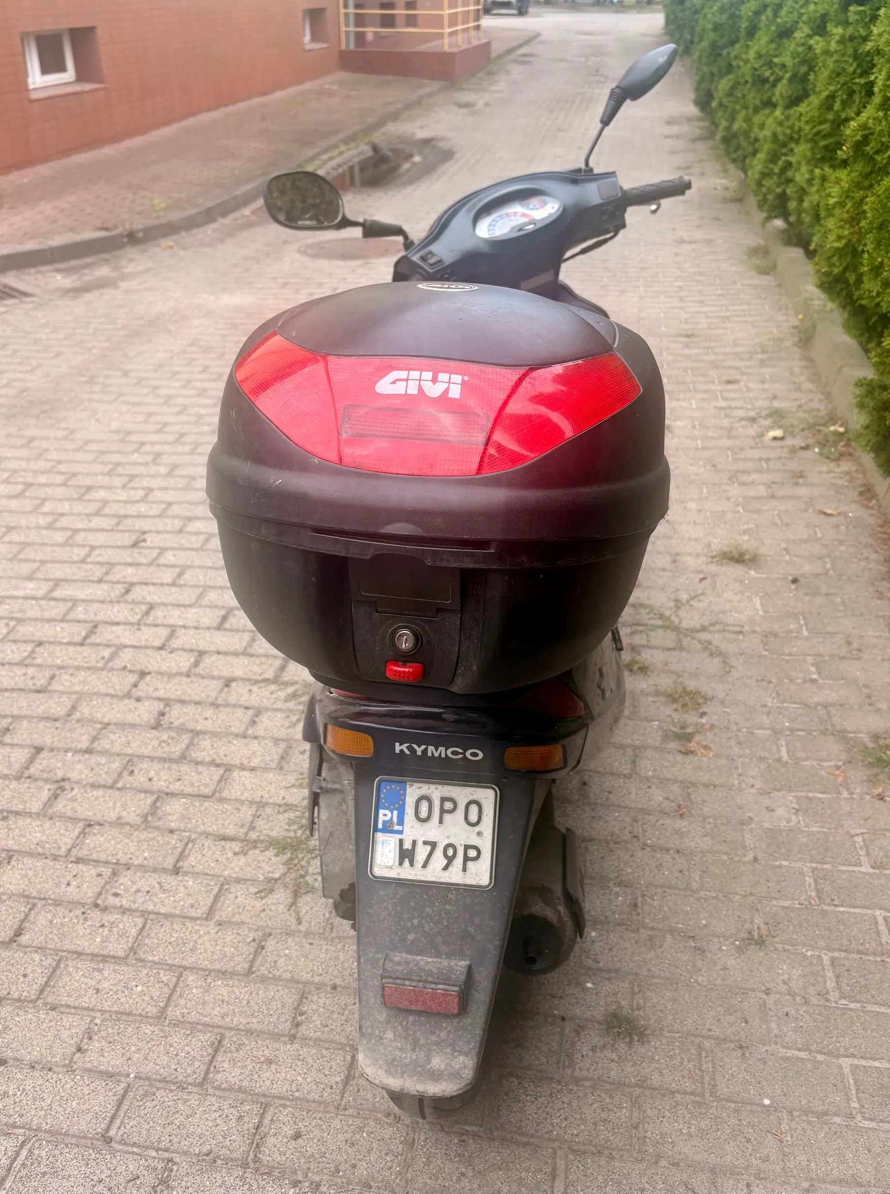 Widok od tyłu na czarny skuter zaparkowany na kostce brukowej. Na tylnym siedzeniu zamontowany jest czarny kufer marki GIVI z czerwonym odblaskiem. Poniżej znajduje się tablica rejestracyjna o numerze OPO W79P z oznaczeniem PL. Nad tablicą widnieje napis KYMCO.