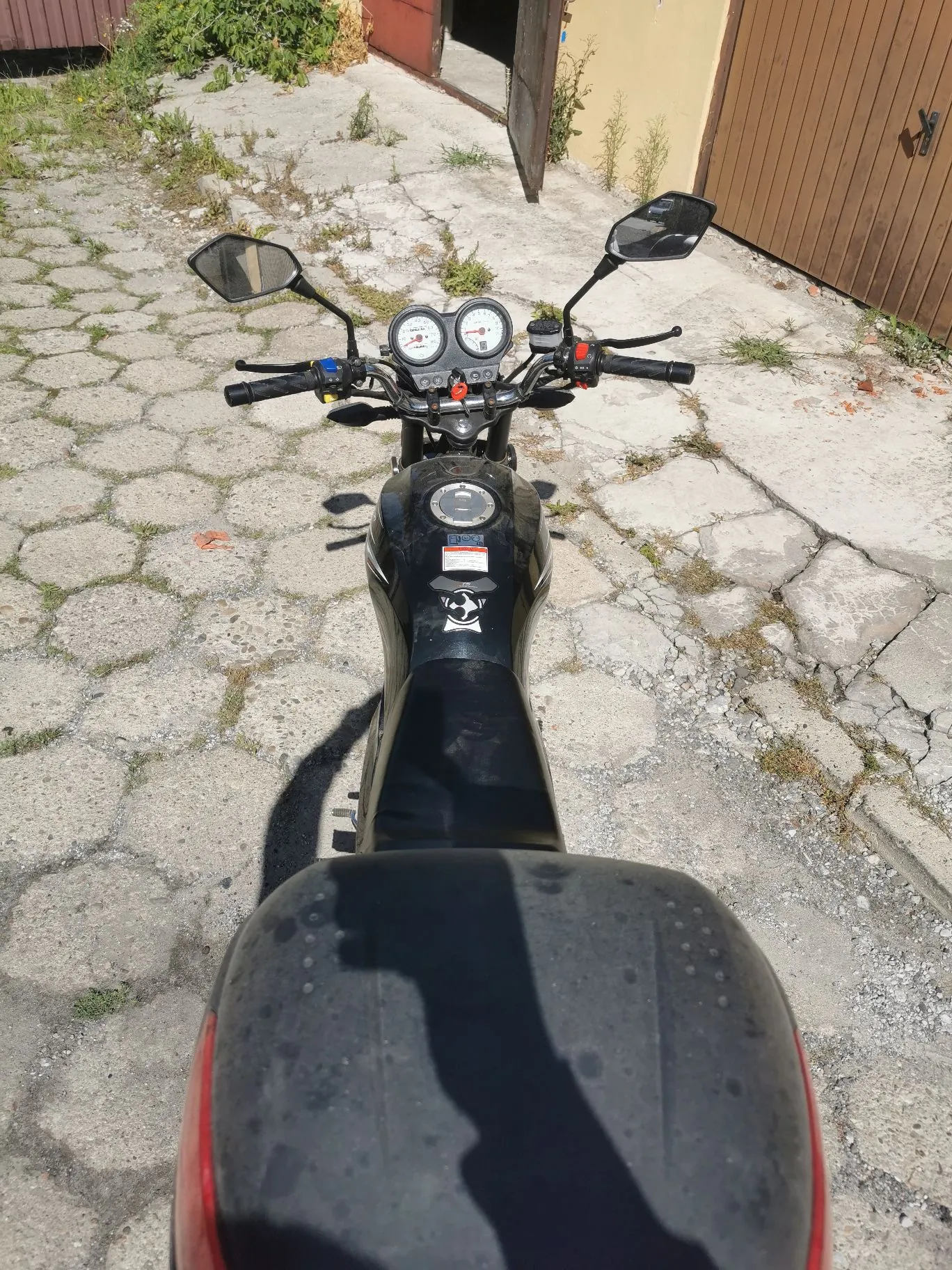 Widok z perspektywy kierowcy na czarny motocykl zaparkowany na betonowym podjeździe z kostki brukowej. Widoczna jest tylna opona, czarne siedzenie, zbiornik paliwa z naklejką oraz kierownica z dwoma okrągłymi zegarami i lusterkami. W tle widać fragmenty budynków i roślinność.
