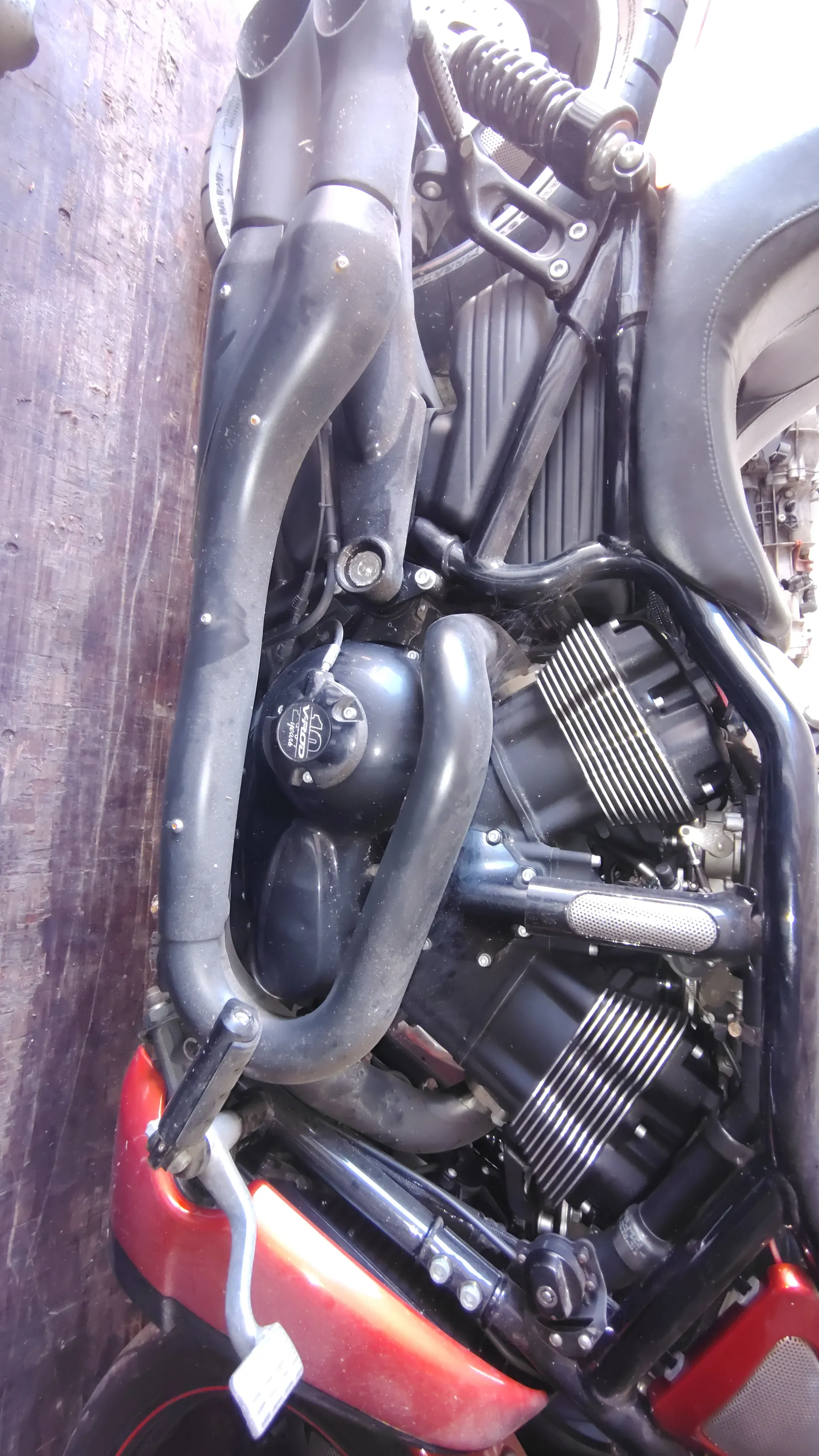 Zbliżenie na silnik motocykla z czarnym siedzeniem, czarnymi i czerwonymi elementami kadłuba, widocznymi rurami wydechowymi oraz chłodzącymi żebrami silnika. Na okrągłym elemencie widoczny napis 'VROD' i '10 years'. Tło stanowi drewniana podłoga oraz części mechaniczne w tle. Czytelny jest również biały etykietka na czerwonym elemencie po prawej stronie.