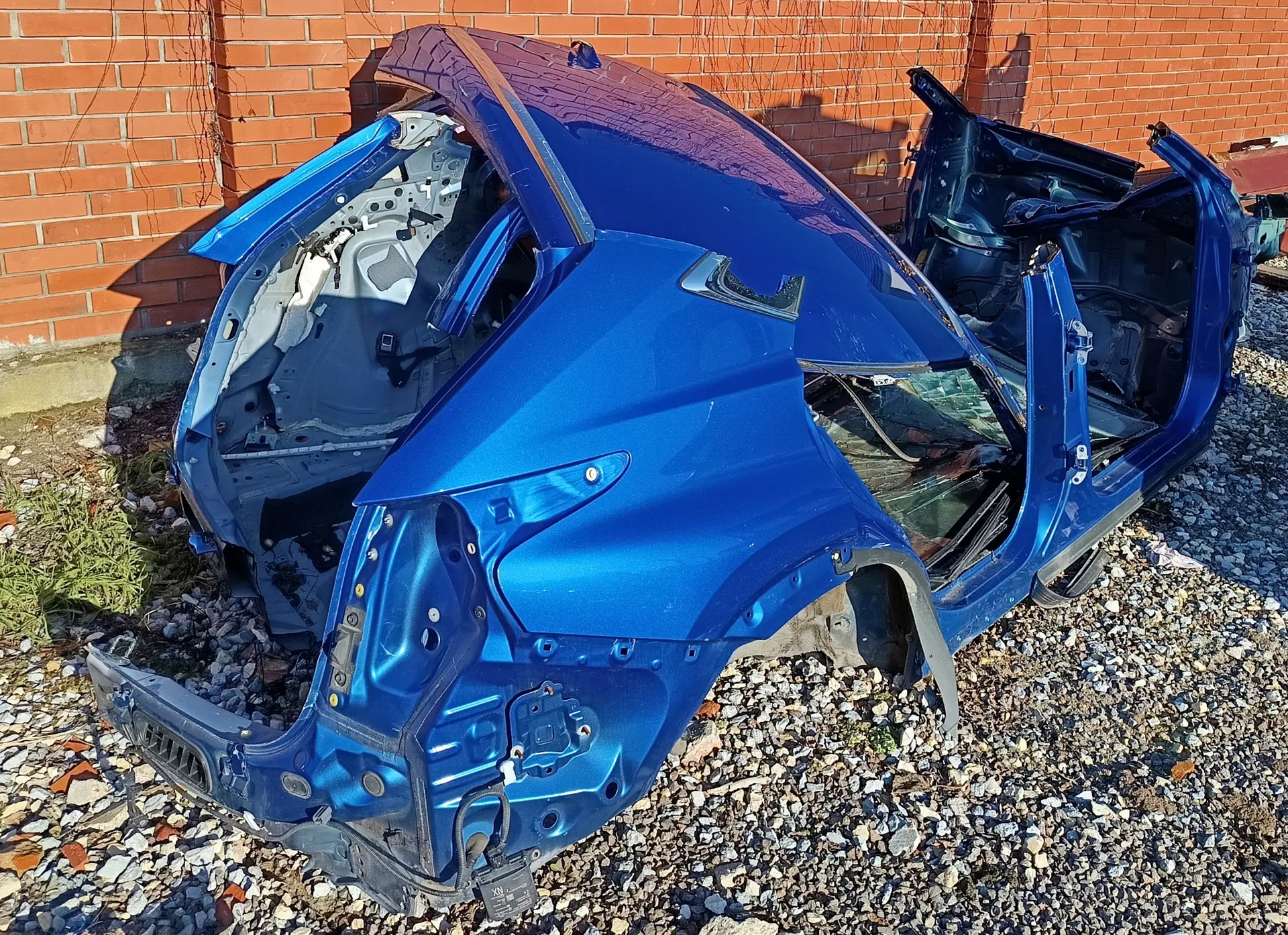 Niebieski karoseria samochodu bez drzwi, maski i dachu, ustawiona na żwirowym podłożu przed ceglaną ścianą. Widoczne są odsłonięte elementy wewnętrzne, w tym fragmenty podłogi i przewodów. W tle ceglana ściana z cieniami, a na ziemi rosną niewielkie łaty trawy. Brak czytelnego tekstu.