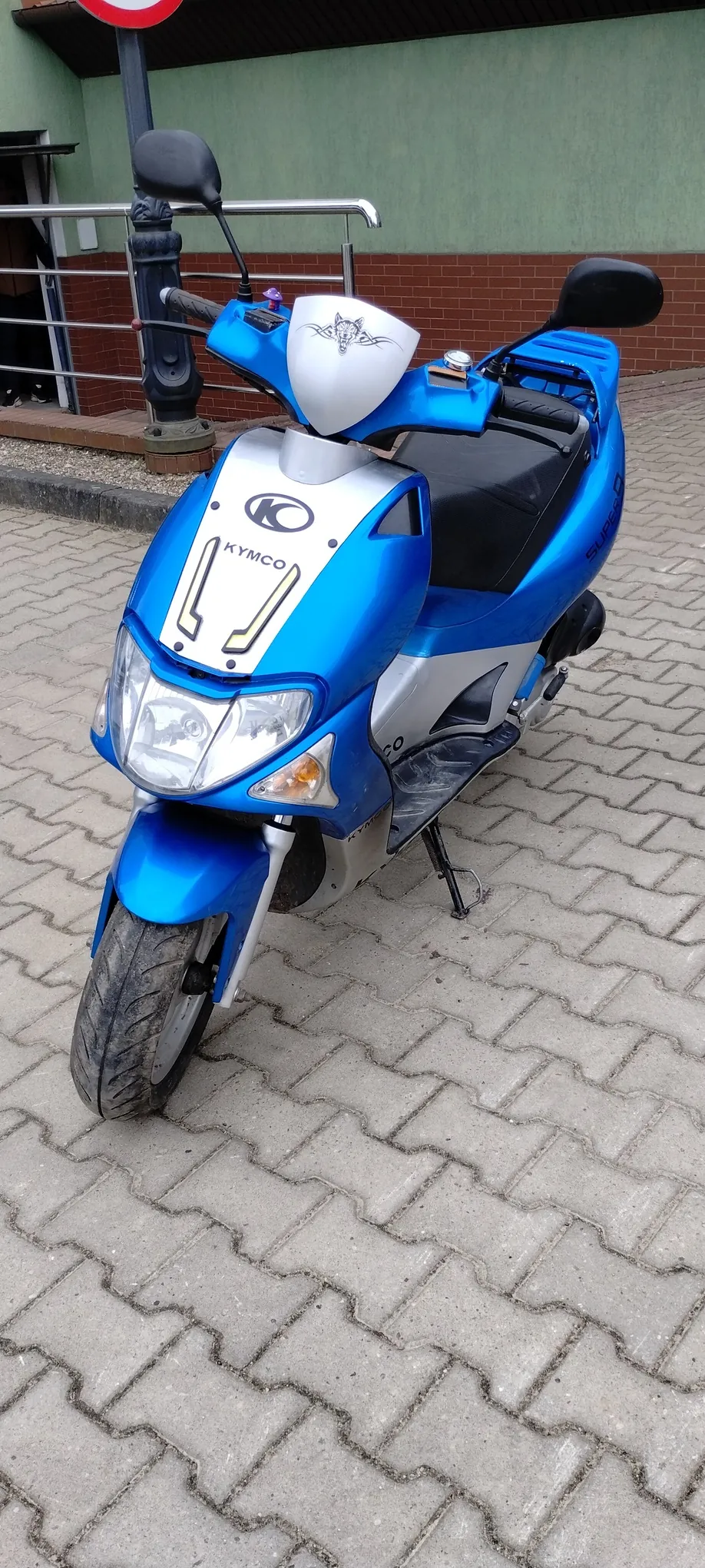 Niebiesko-biały skuter marki Kymco zaparkowany na chodniku z kostki brukowej. Widoczne są logo Kymco na przodzie, boczne lusterka, reflektory oraz fragment budynku z zieloną ścianą i ceglaną elewacją. W lewym dolnym rogu częściowo widoczny znak drogowy z czerwonym obramowaniem.