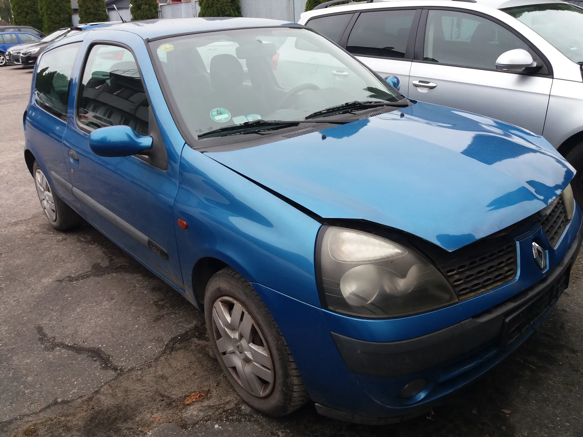 Niebieski trzydrzwiowy samochód osobowy marki Renault z widocznym uszkodzeniem na masce, zaparkowany na placu parkingowym. Na przedniej szybie znajdują się naklejki, w tym zielona okrągła. W tle widoczne są inne pojazdy, budynki oraz drzewa. Samochód posiada srebrne felgi i czarne zderzaki.