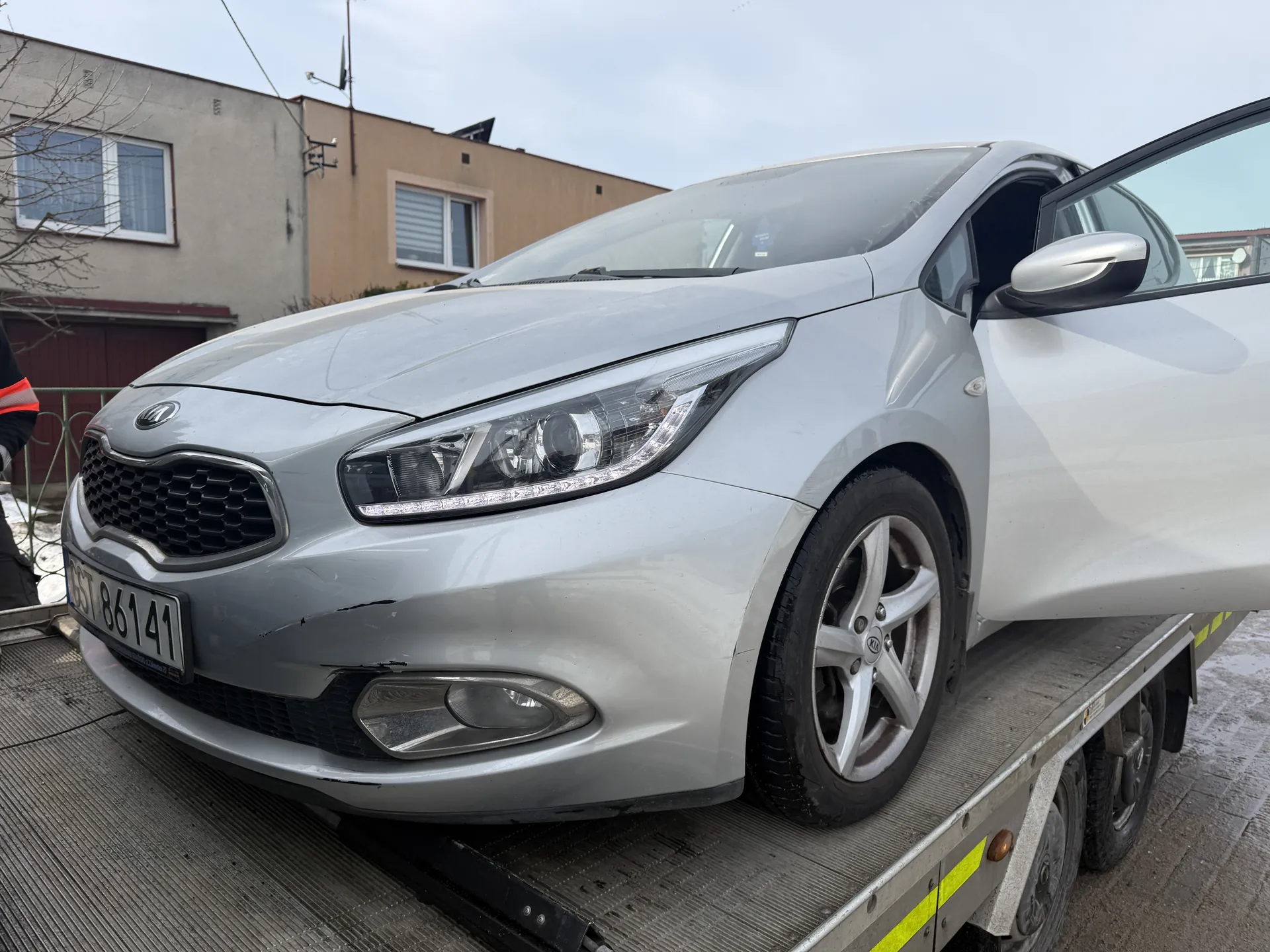 Srebrny samochód marki Kia z otwartą przednią prawą drzwiami, stojący na platformie holownika. Widoczna tablica rejestracyjna CST 86141, uszkodzony przedni zderzak oraz reflektory. W tle budynki mieszkalne, bezlistne drzewo i osoba w czarno-czerwonym ubraniu. Podłoże pokryte jest śniegiem i lodem.