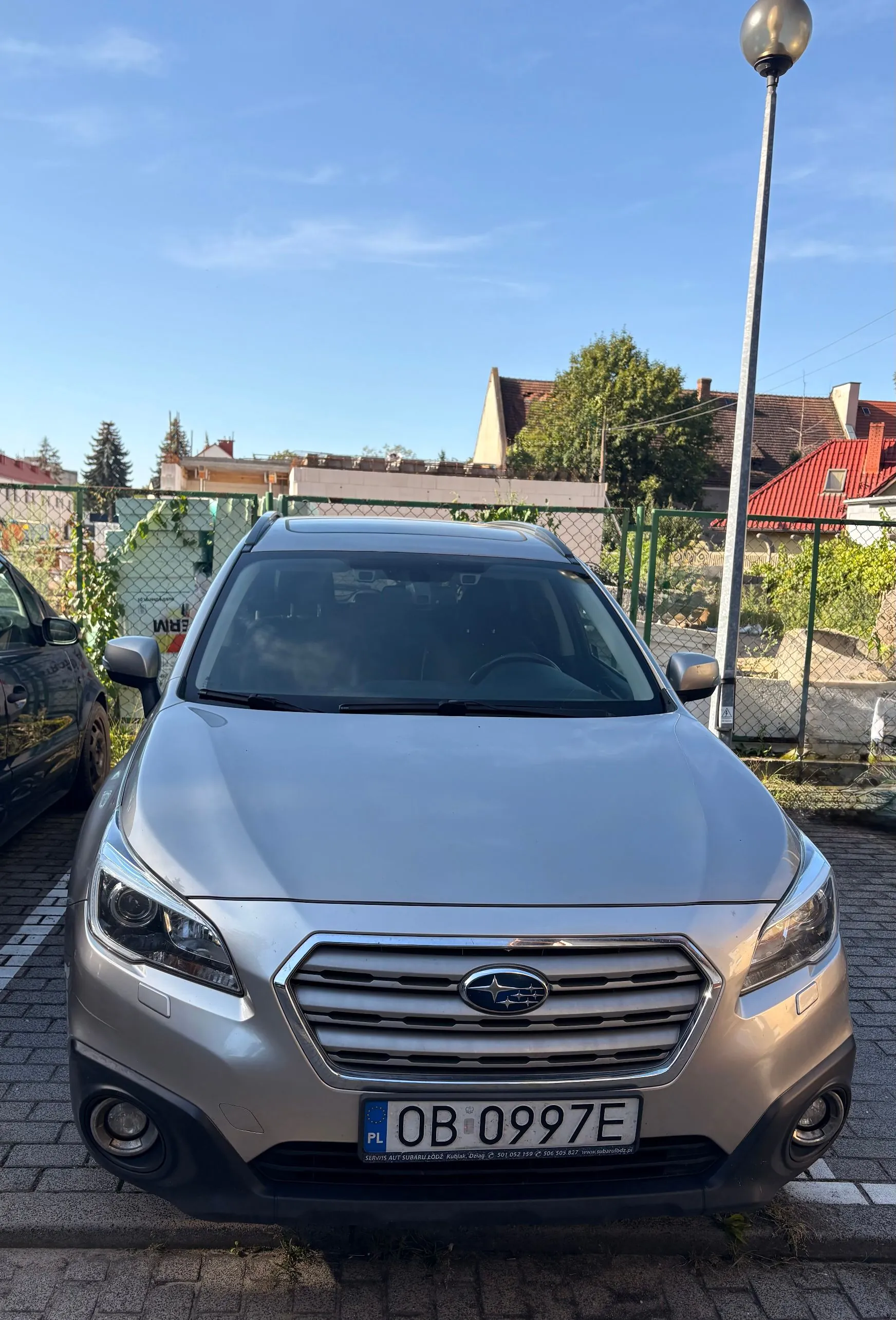 Srebrny samochód marki Subaru zaparkowany przodem do obiektywu. Widoczna jest tablica rejestracyjna o numerze OB 0997E z oznaczeniem PL. W tle znajduje się zielony płot z siatki, drzewa oraz dachy budynków pod jasnoniebieskim niebem. Po prawej stronie widoczna jest wysoka latarnia uliczna.