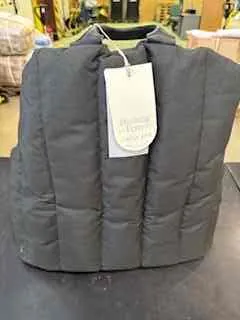 Czarna pikowana kamizelka leżąca na ciemnej powierzchni. Na kamizelce widoczny biały etykietka z nieczytelnym tekstem. W tle rozmyty widok wnętrza sklepu z półkami i przedmiotami.