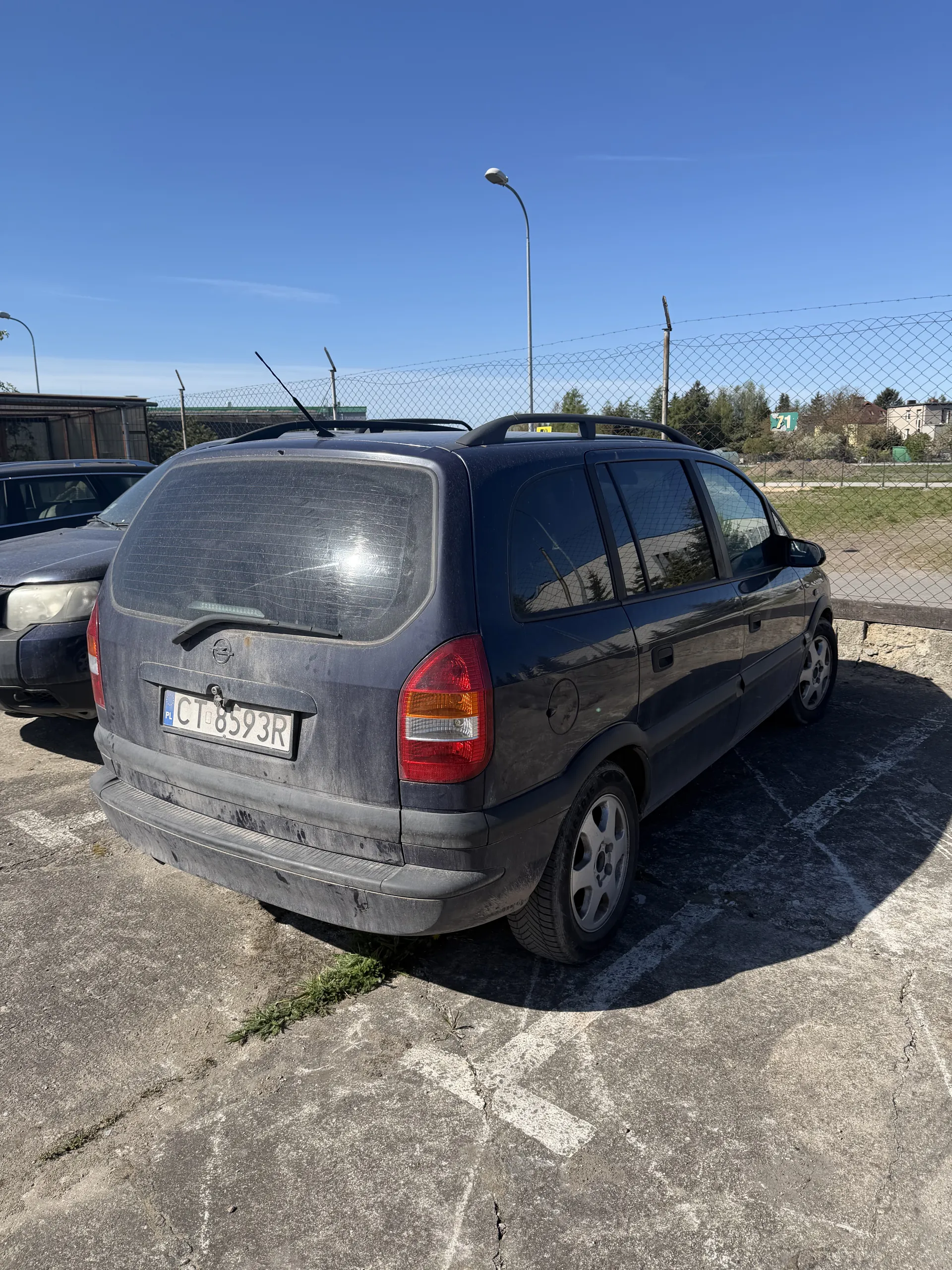 Ciemny samochód osobowy zaparkowany na asfaltowym placu parkingowym. Widoczna tablica rejestracyjna CT8593R. W tle siatka ogrodzeniowa, trawa, drzewa, latarnia uliczna oraz błękitne niebo bez chmur. Samochód ma zabrudzoną karoserię i dachowy bagażnik.