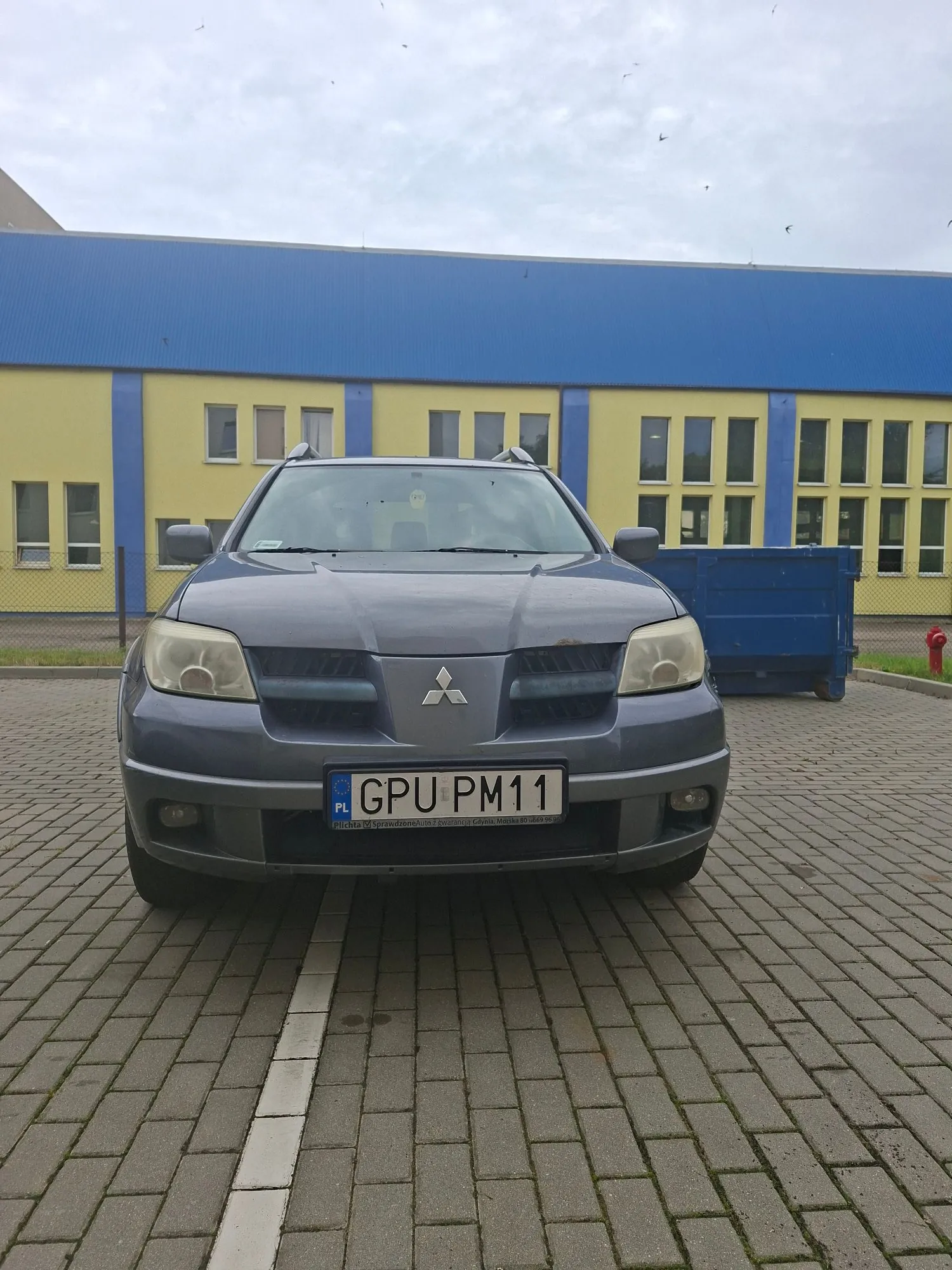 Srebrny samochód marki Mitsubishi zaparkowany na kostce brukowej przed żółto-niebieskim budynkiem. Samochód ma tablicę rejestracyjną o numerze GPU PM11. Obraz jest zorientowany poziomo, mimo że scena powinna być pionowa.