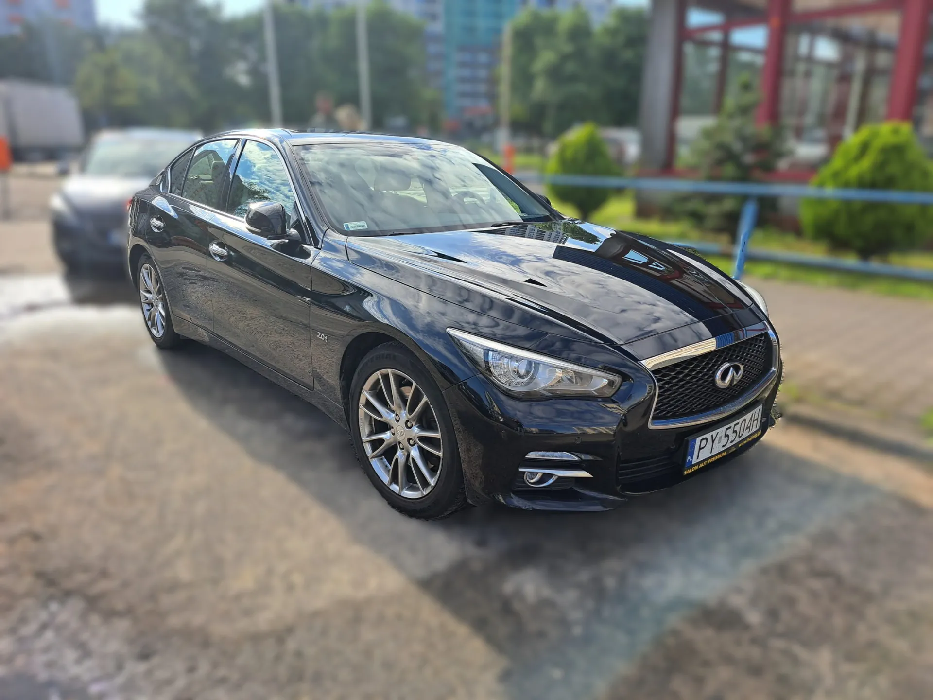 Czarny samochód marki Infiniti zaparkowany na mokrym podłożu. Pojazd jest widoczny z przodu i z boku, z tablicą rejestracyjną o numerze PY 5504H. W tle widoczne są rozmyte drzewa, budynki oraz niebieska barierka.