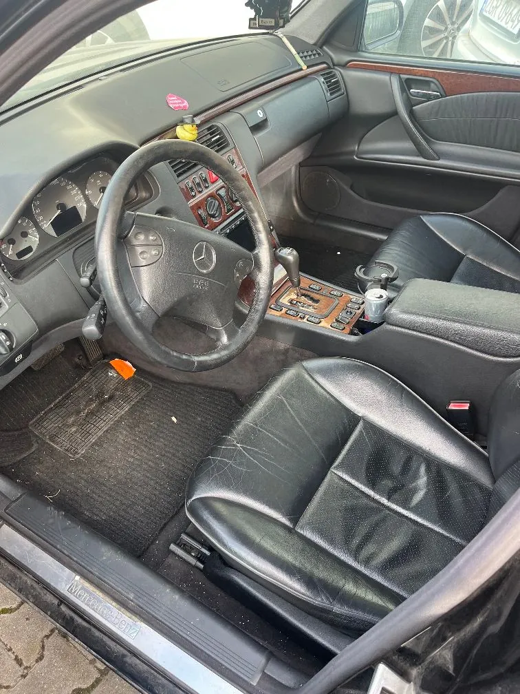 Wnętrze samochodu marki Mercedes-Benz z widokiem na stronę kierowcy. Widoczna jest czarna skórzana tapicerka foteli, czarna kierownica z logo Mercedesa, deska rozdzielcza z elementami drewnianymi oraz zegary w kokpicie. Na progu samochodu widoczny jest napis 'Mercedes-Benz'.