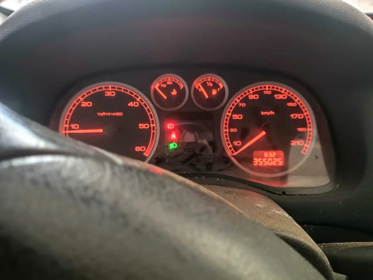 Zbliżenie na deski rozdzielcze samochodu z czerwonym podświetleniem. Po lewej stronie wskaźnik obrotomieru (t/min x100) z zakresem do 60, po prawej prędkościomierz (km/h) z zakresem do 210. W środku dwa mniejsze wskaźniki oraz czerwone i zielone lampki ostrzegawcze. Na wyświetlaczu cyfrowym widoczny przebieg 355025 oraz wartość 832. W dolnej części kadru fragment kierownicy.