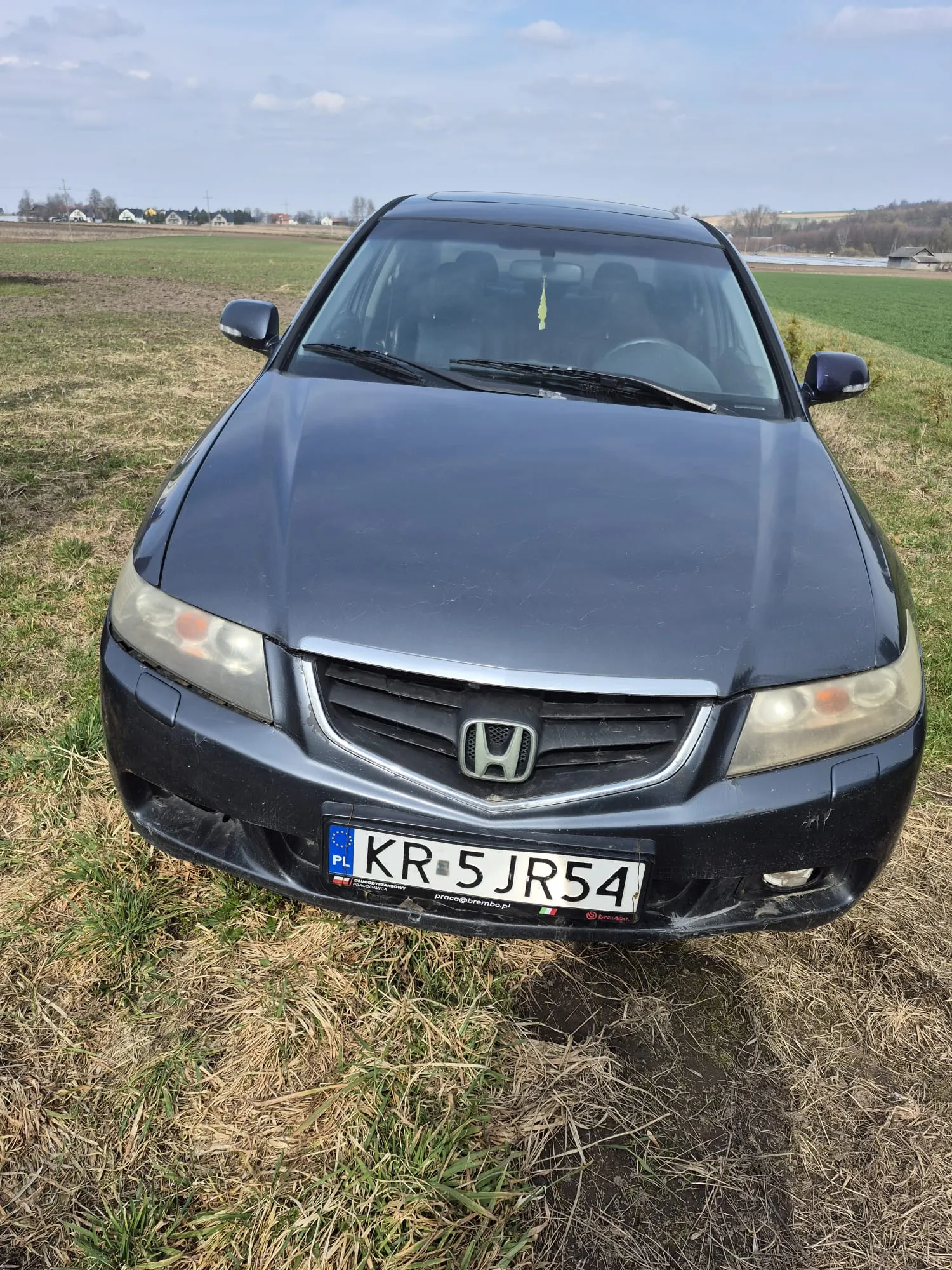 Ciemnoszary samochód marki Honda z tablicą rejestracyjną KR 5 JR54, stojący na trawiastym polu. Na tablicy widoczny napis PL oraz dodatkowy napis praca@brembo.pl. W tle rozległe pola, domy i częściowo zachmurzone niebo. Na przedniej szybie wiszący żółty przedmiot.