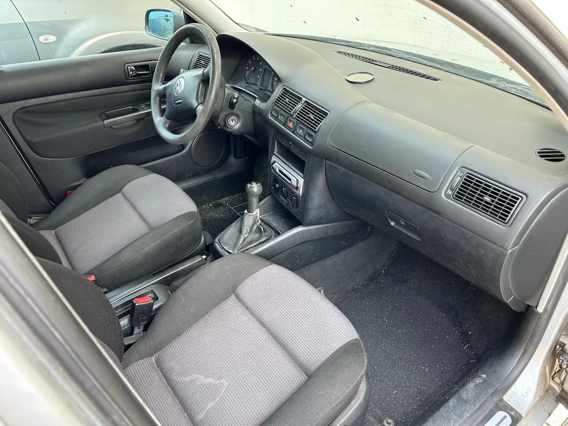 Wnętrze samochodu z perspektywy otwartych drzwi pasażera. Widoczna jest czarna deska rozdzielcza, kierownica z logo Volkswagena, panel instrumentów, konsola środkowa z radiem i lewarkiem zmiany biegów oraz dwa przednie fotele z szarym materiałem w centralnej części.