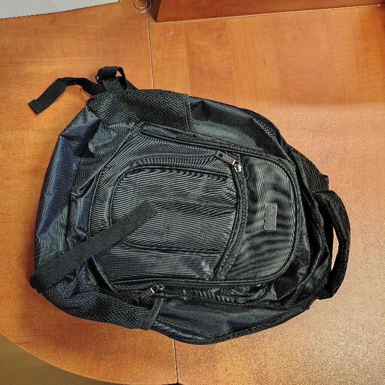 Czarna, materiałowa torba sportowa lub plecak leżący na brązowym, drewnianym blacie. Torba posiada kilka zamków błyskawicznych, przednią kieszeń oraz regulowany pasek.
