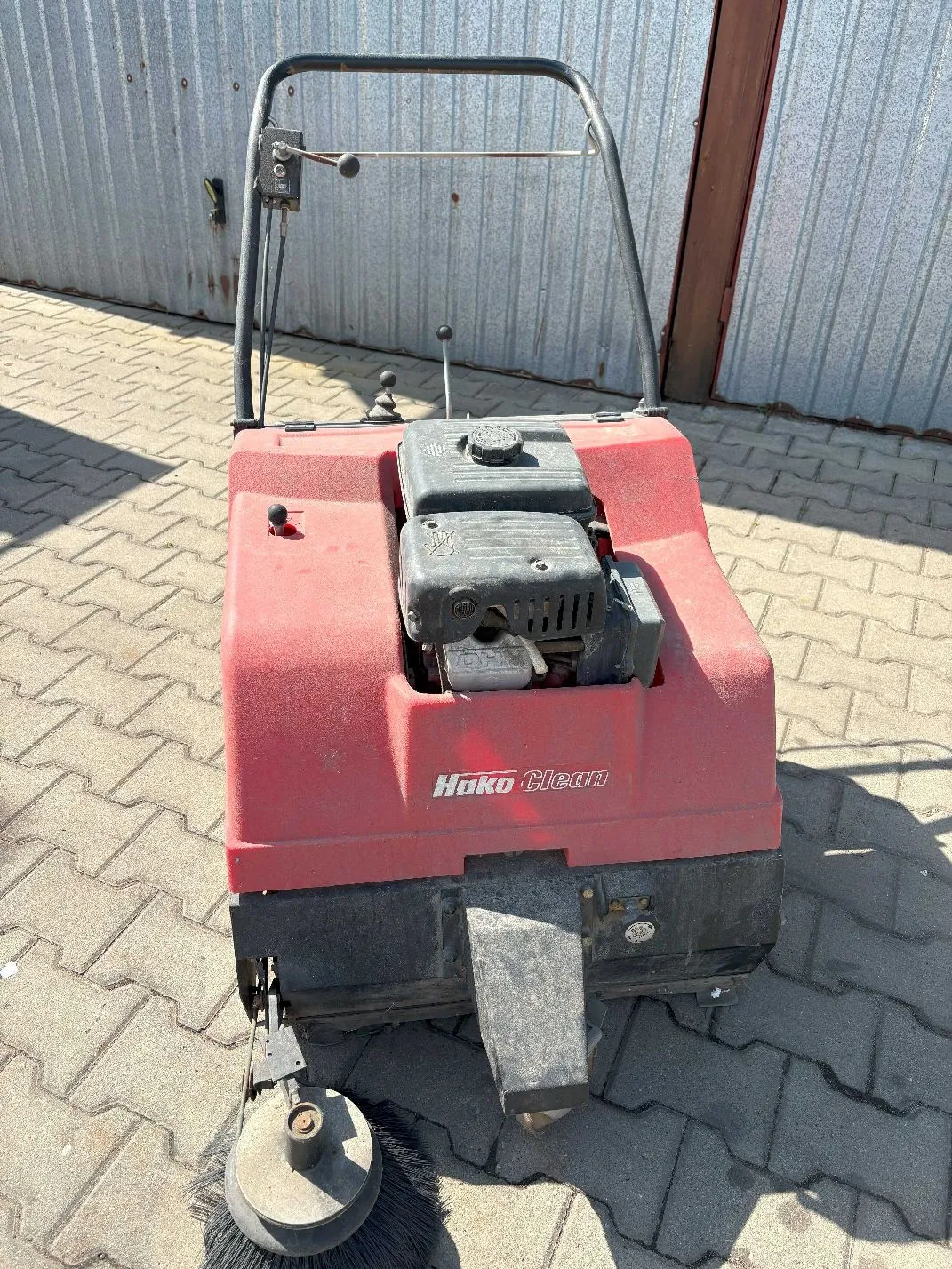 Czerwona maszyna czyszcząca marki Hako Clean z silnikiem spalinowym, ustawiona na kostce brukowej przed metalową ścianą. Urządzenie posiada czarną rączkę sterującą oraz szczotkę obrotową z przodu.