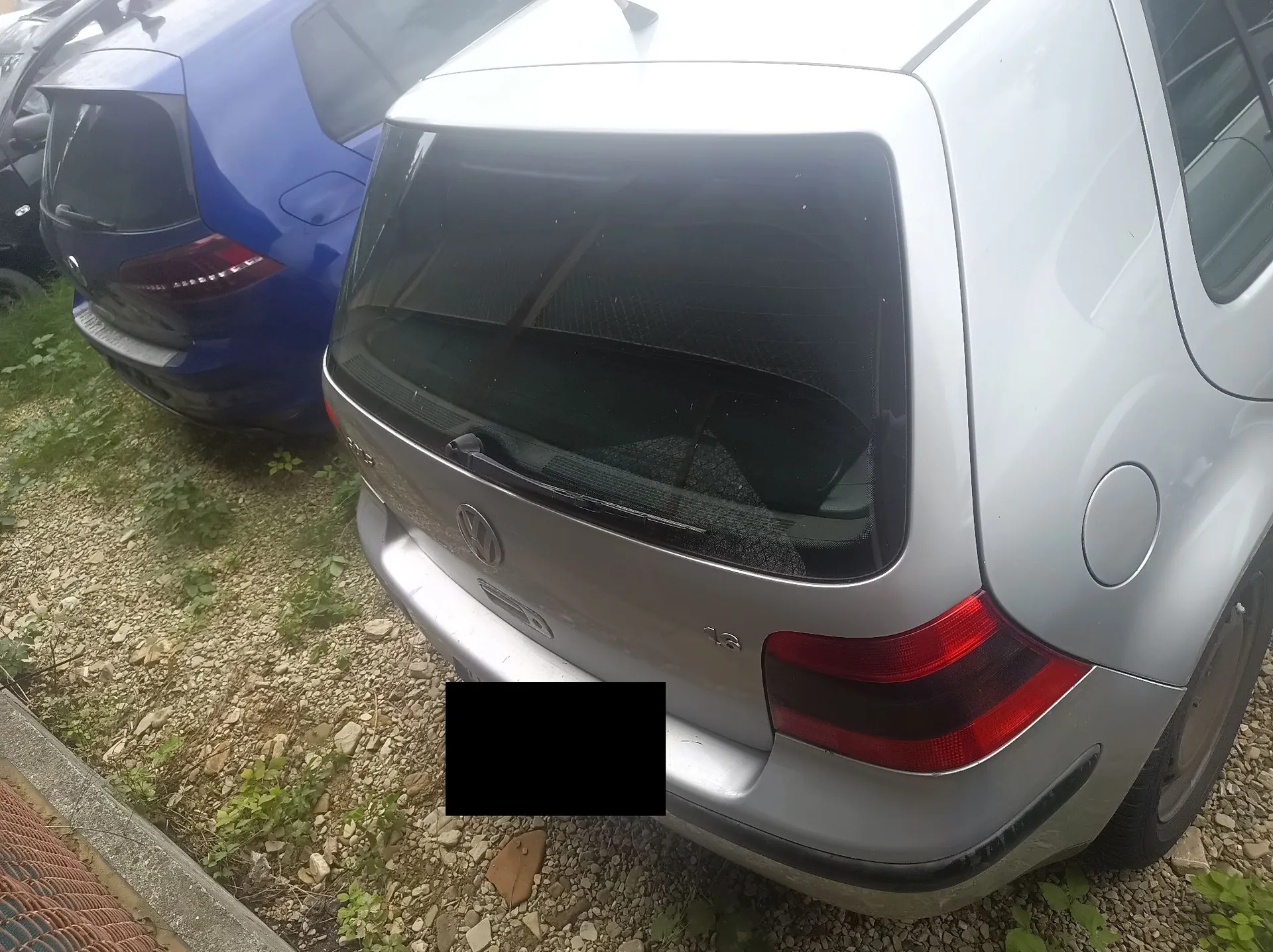 Tylna część srebrnego samochodu marki Volkswagen zaparkowanego obok niebieskiego samochodu. Widoczna jest tylna szyba, logo Volkswagena oraz prawe tylne światło. Tablica rejestracyjna jest zakryta czarnym prostokątem. Samochody stoją na żwirowym podłożu z kępkami zielonej trawy.