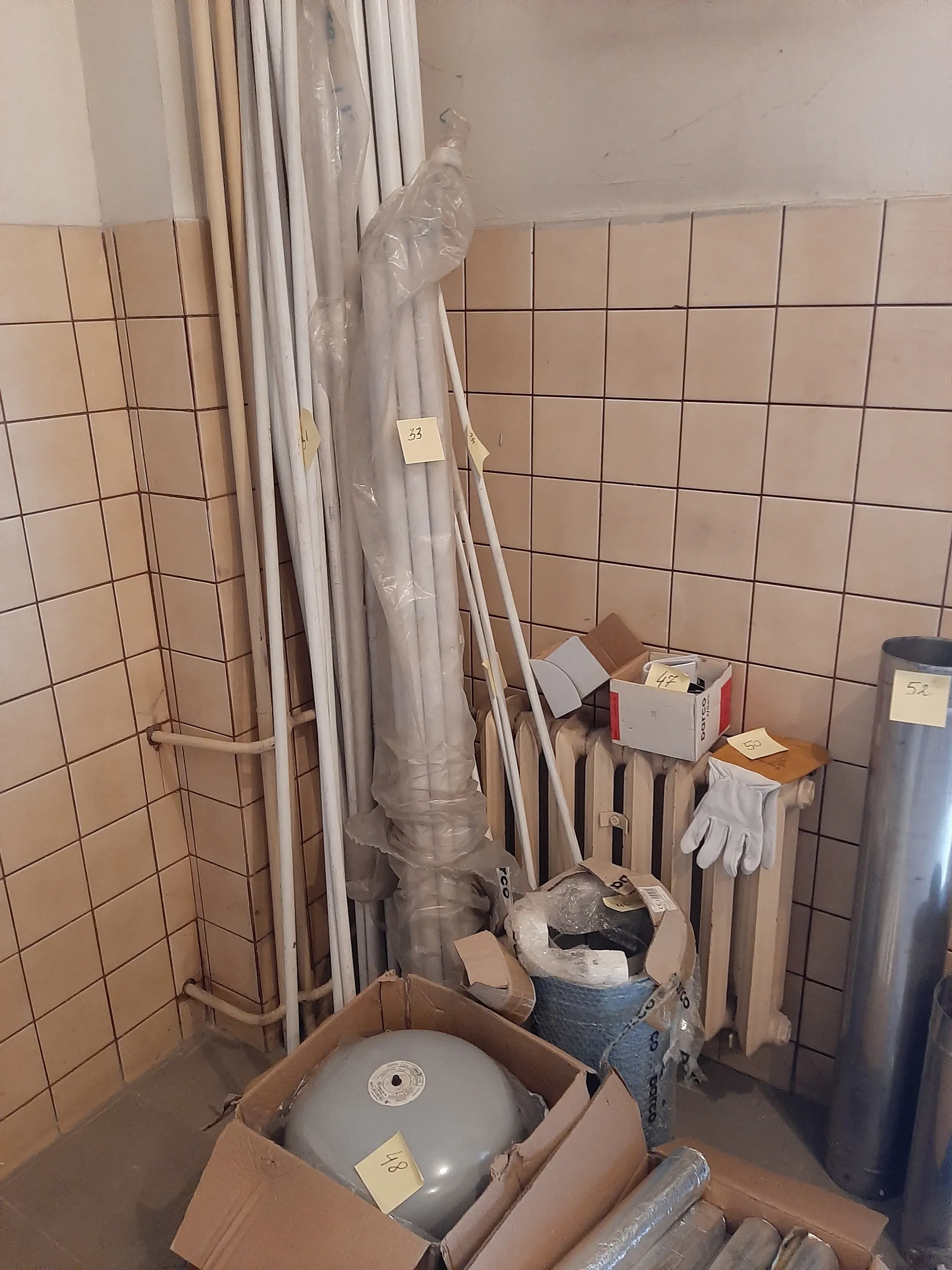 Pomieszczenie z beżowymi kwadratowymi kafelkami na ścianie. W centralnej części leżą długie białe rury owinięte przezroczystą folią, oznaczone żółtymi etykietami z numerami (45, 47, 48). W prawym górnym rogu znajdują się pudełka kartonowe, w jednym z nich szary okrągły zbiornik z etykietą 48. Na białym grzejniku leżą białe rękawice robocze oraz pudełko z widocznym napisem BOSCH. Nad grzejnikiem umieszczona jest metalowa rura. W tle widoczne są dodatkowe materiały budowlane i opakowania.