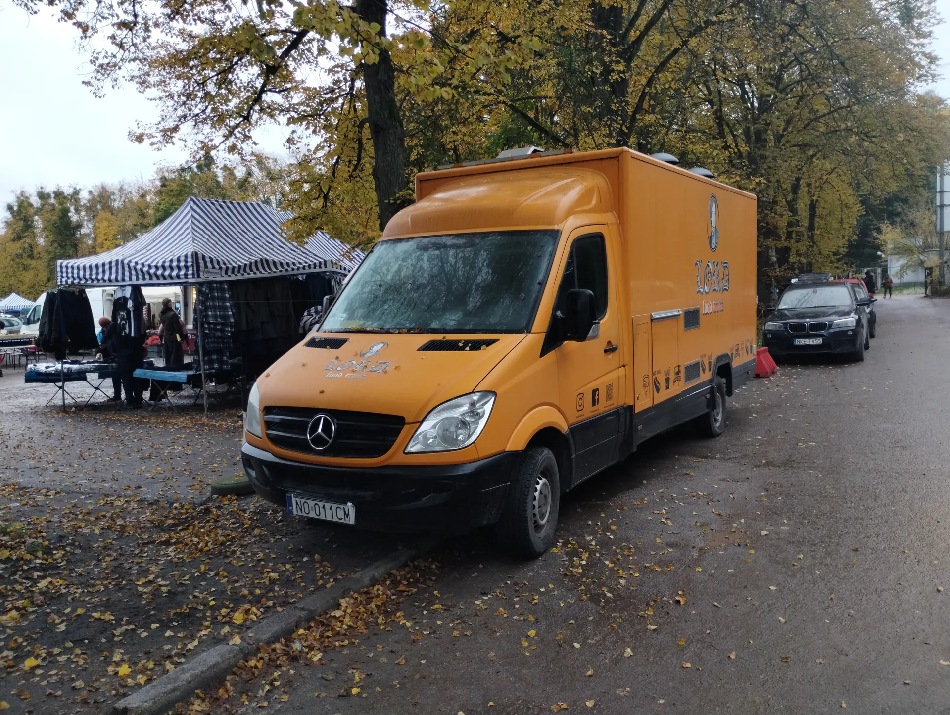 Pomarańczowy furgon Mercedes-Benz z napisem "LOID food truck" na boku i przodzie, tablicą rejestracyjną "NO 011CM". Za furgonem czarny samochód osobowy. Po lewej stronie namiot w paski z ubraniami na wieszakach i ludźmi. Tło z drzewami o żółtych liściach, opadłymi liśćmi na ziemi i pochmurnym niebem.
