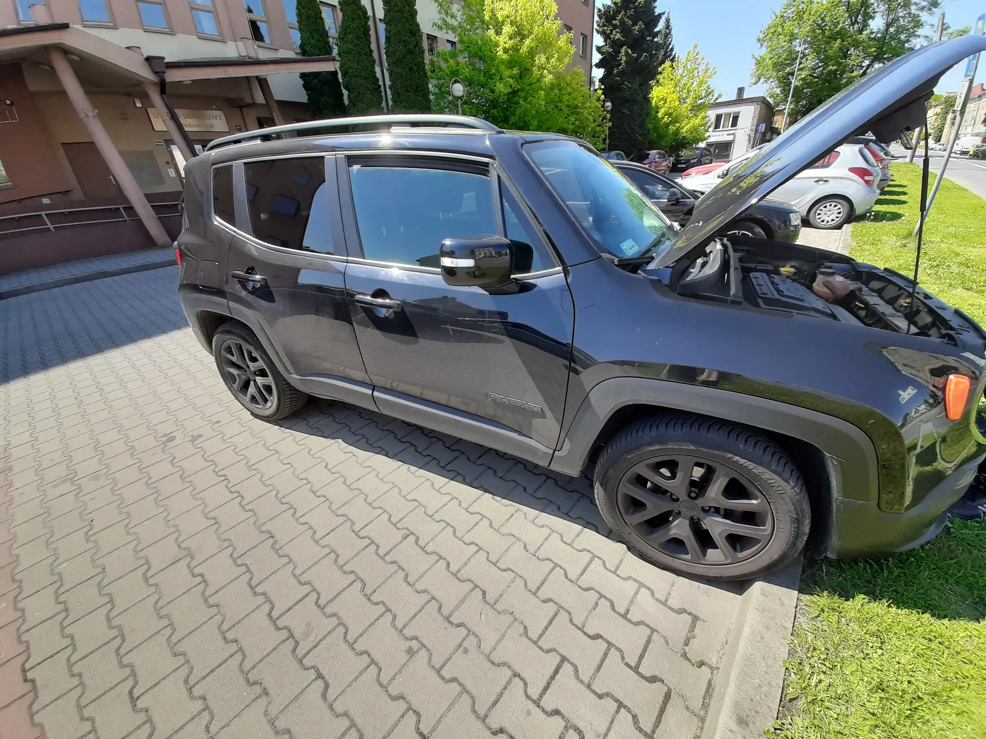 Ciemnoszary SUV typu Renegade z otwartą maską zaparkowany na brukowanej powierzchni obok trawnika. W tle widoczne są budynki, drzewa, inne zaparkowane samochody oraz jasne niebo. Na boku pojazdu widnieje napis "Renegade". Samochód stoi na chodniku z płytek w geometrycznym wzorze, a w tle znajduje się budynek z napisem na szyldzie.