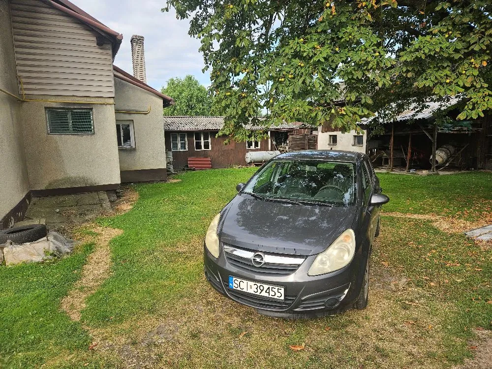 Szary samochód marki Opel zaparkowany na trawniku przed niskimi, gospodarczymi budynkami. Samochód ma polską tablicę rejestracyjną o numerze SCI 39455. W tle widoczne są drewniane zabudowania, komin oraz duże drzewo z zielonymi liśćmi zwisającymi nad pojazdem.