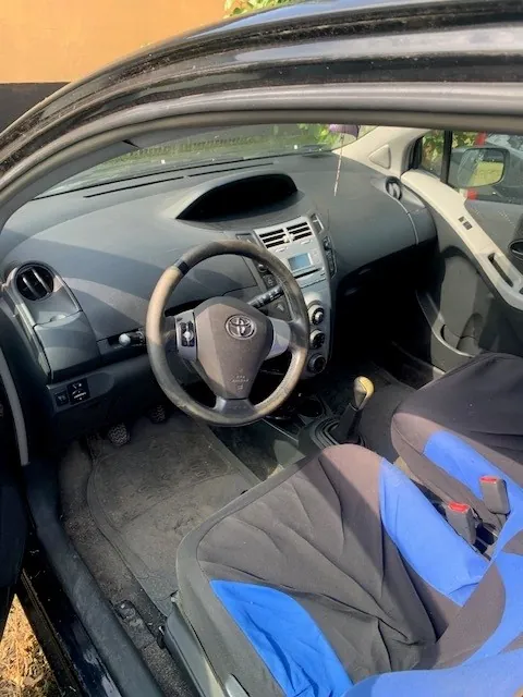 Wnętrze samochodu marki Toyota widziane z perspektywy otwartych drzwi kierowcy. Widoczna jest czarna kierownica z logo Toyoty, deska rozdzielcza z systemem audio i nawiewami, lewarek zmiany biegów oraz dwa fotele z czarno-niebieskimi pokrowcami. Na podłodze znajduje się ciemna mata samochodowa.