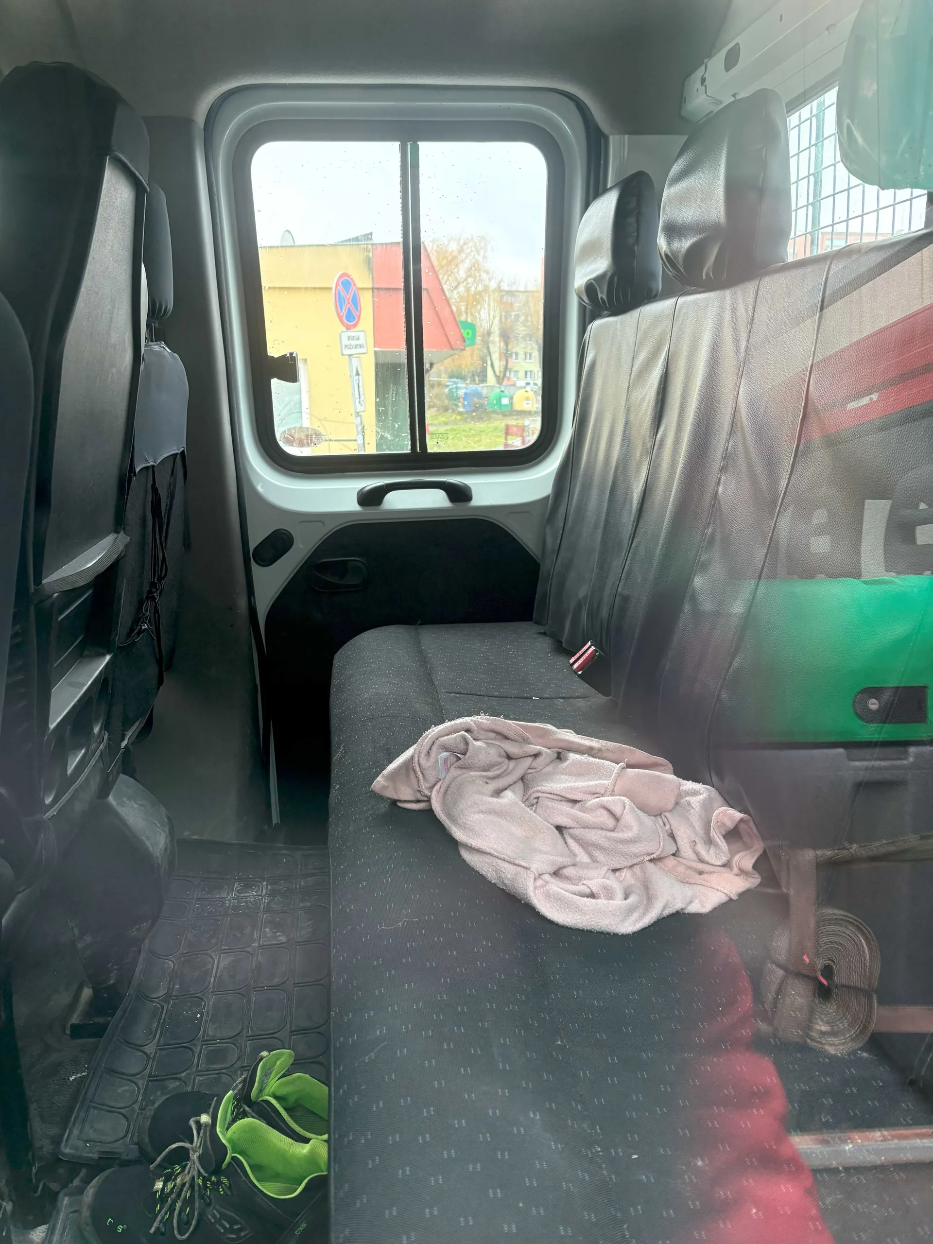 Wnętrze pojazdu z czarnymi siedzeniami, na jednym z nich leży różowy ręcznik. Na podłodze widoczne czarne buty z zielonymi elementami. Przez tylne okno widać żółty budynek z czerwonym dachem, znak zakazu postoju (niebieski okrąg z czerwonym krzyżem) oraz zielone pojemniki na śmieci. Na zewnątrz jest pochmurno, a na szybie widoczne są krople deszczu.