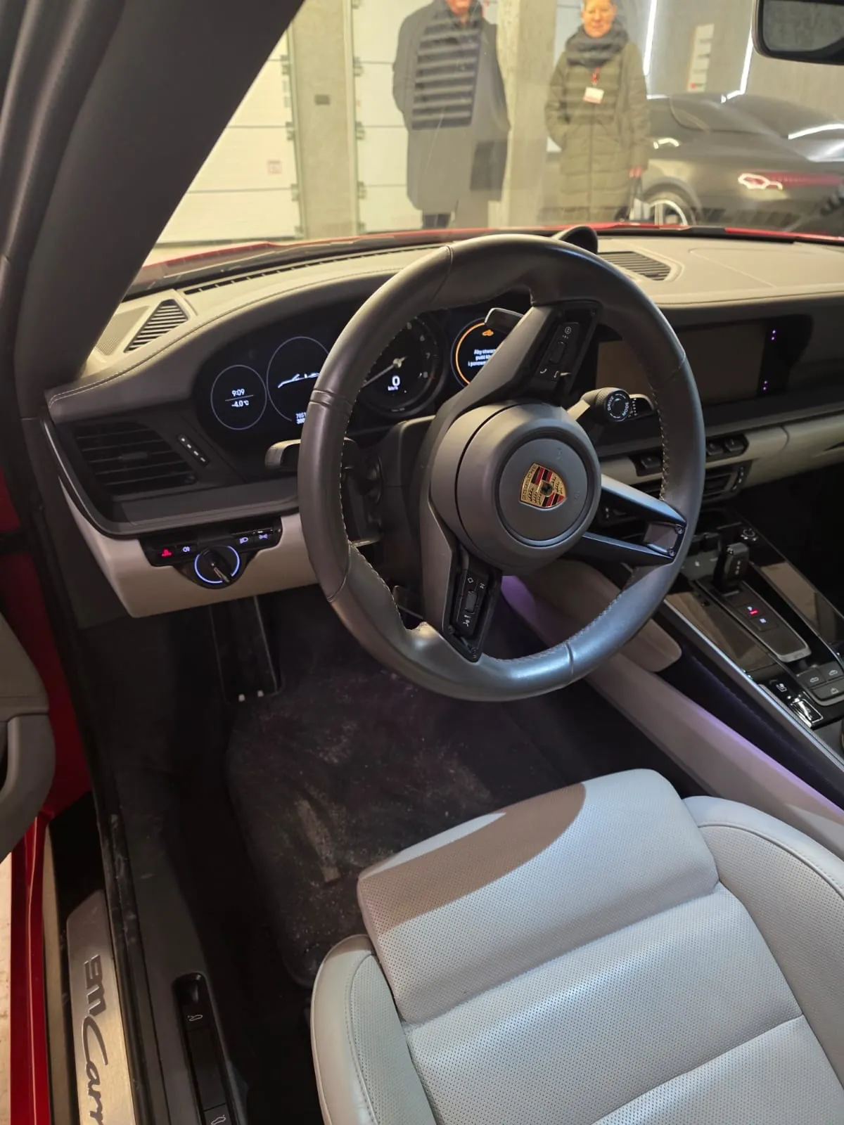 Wnętrze samochodu Porsche z widocznym kierownicą z logo marki, cyfrowym zegarem pokazującym godzinę 9:09 i temperaturę -4°C, jasnymi skórzonymi siedzeniami oraz fragmentem deski rozdzielczej. Na zewnątrz przez przednią szybę widać dwóch stojących osób i inny samochód. Na progu drzwi widoczny napis Cayman.