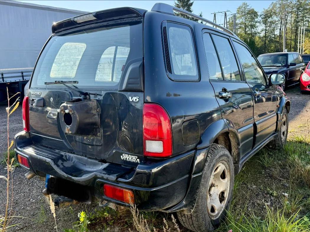 Czarny samochód terenowy marki Suzuki Grand Vitara V6 widziany od tyłu i z boku. Pojazd posiada zamontowany na klapie bagażnika uchwyt na koło zapasowe (bez koła), czarne relingi dachowe oraz srebrne felgi aluminiowe. Na tylnej klapie widoczne są napisy 'SUZUKI', 'V6' oraz 'GRAND VITARA'. Samochód stoi na nieutwardzonym terenie z trawą, w tle widoczny jest fragment białej naczepy ciężarówki oraz inne zaparkowane samochody.