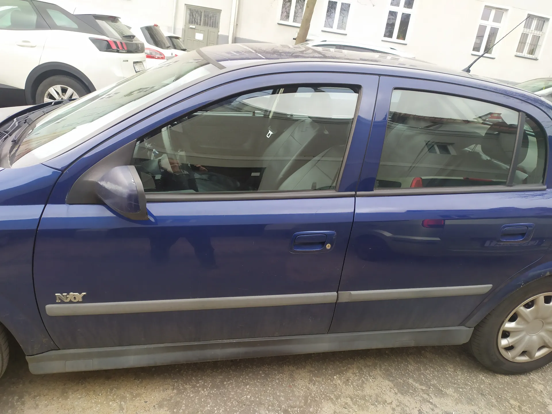 Boczny widok niebieskiego samochodu osobowego marki Nissan z napisem Njoy na przedniej fenderze. Samochód stoi na podwórku z żwirem, w tle widoczne są inne pojazdy (biały SUV i czerwony samochód) oraz budynek z oknami i drzwiami. Wnętrze samochodu pokazuje siedzenia i kierownicę. Na boku pojazdu widoczne są klamki drzwi i lusterko boczne.