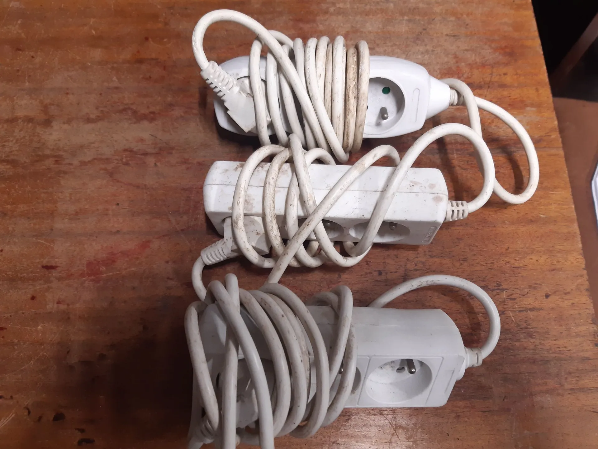 Trzy białe przedłużacze elektryczne leżące na drewnianym blacie. Każdy z nich ma zwinięty biały kabel. Górny i dolny przedłużacz to pojedyncze gniazdka, natomiast środkowy jest listwą z trzema gniazdami. Przedłużacze są lekko zabrudzone.