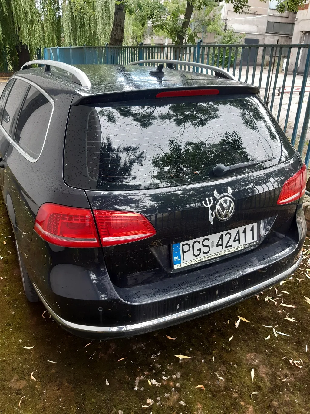 Tylny widok czarnego samochodu marki Volkswagen w nadwoziu kombi. Pojazd posiada srebrne relingi dachowe i polską tablicę rejestracyjną o numerze PGS 42411. Nad logo Volkswagena znajduje się biała naklejka przedstawiająca trójząb. Samochód stoi na ciemnym, wilgotnym podłożu z opadłymi liśćmi, w tle widoczny jest niebieski metalowy płot oraz zielone drzewa.