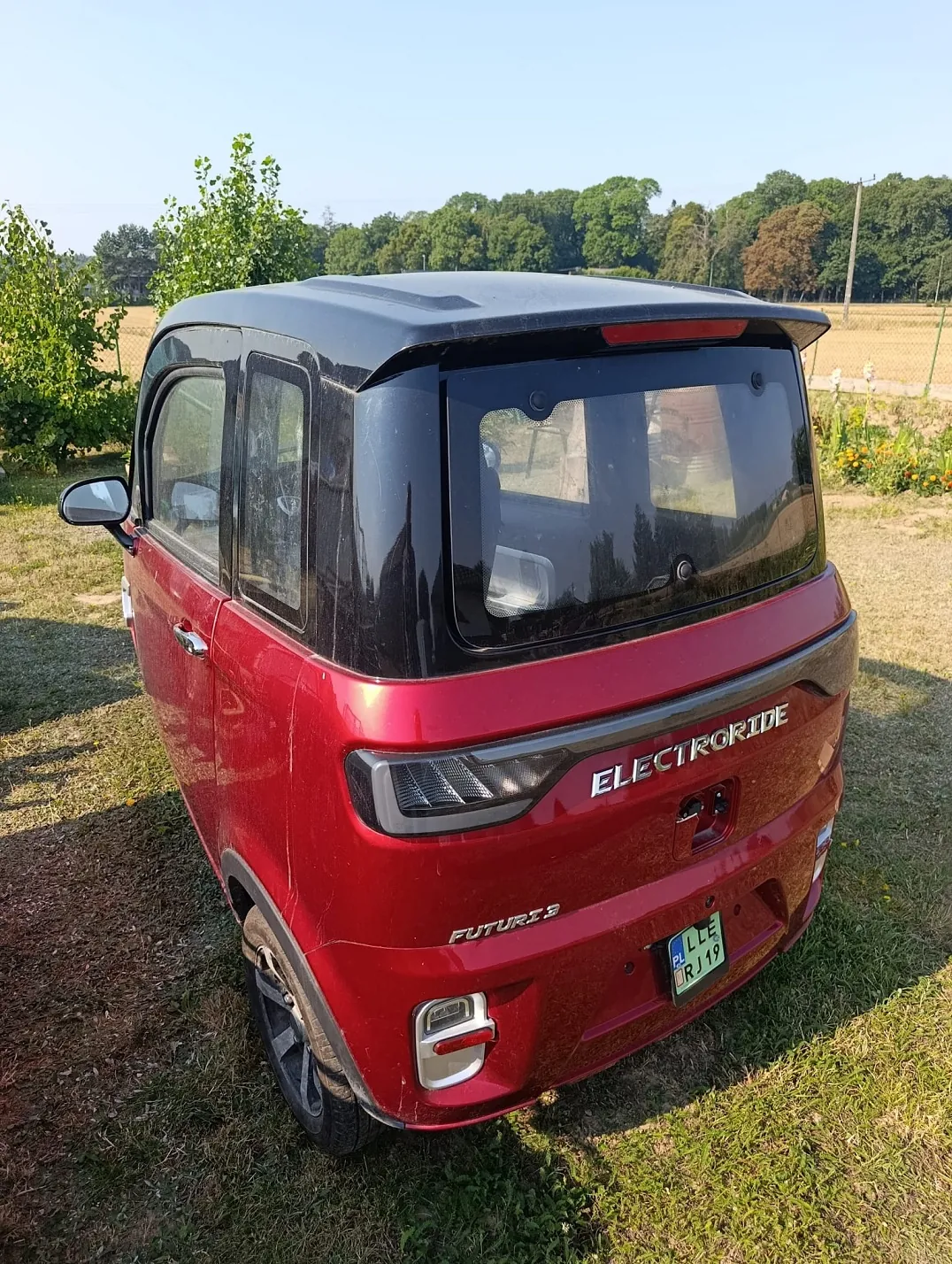 Czerwony, kompaktowy samochód elektryczny marki ElectroRide, model Future 3, zaparkowany na trawiastym terenie. Pojazd ma czarny dach i tylną szybę. Na tylnej klapie widnieje napis 'ELECTRORIDE', a poniżej mniejszy napis 'FUTURE 3'. Samochód posiada tablicę rejestracyjną o numerze 'LLE RJ 19'. W tle widać zielone drzewa i pole pod jasnym niebem.