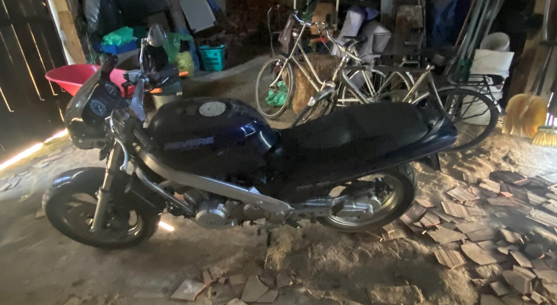 Ciemnoniebieski motocykl z napisem 'REVERSE' na zbiorniku paliwa, stojący w garażu lub pomieszczeniu magazynowym. W tle widoczne są dwa rowery, czerwony zlew, zielone pojemniki, narzędzia oraz rozrzucone na podłodze kawałki płytek. Podłoga jest brudna, a w tle znajdują się różne przedmioty gospodarstwa domowego.