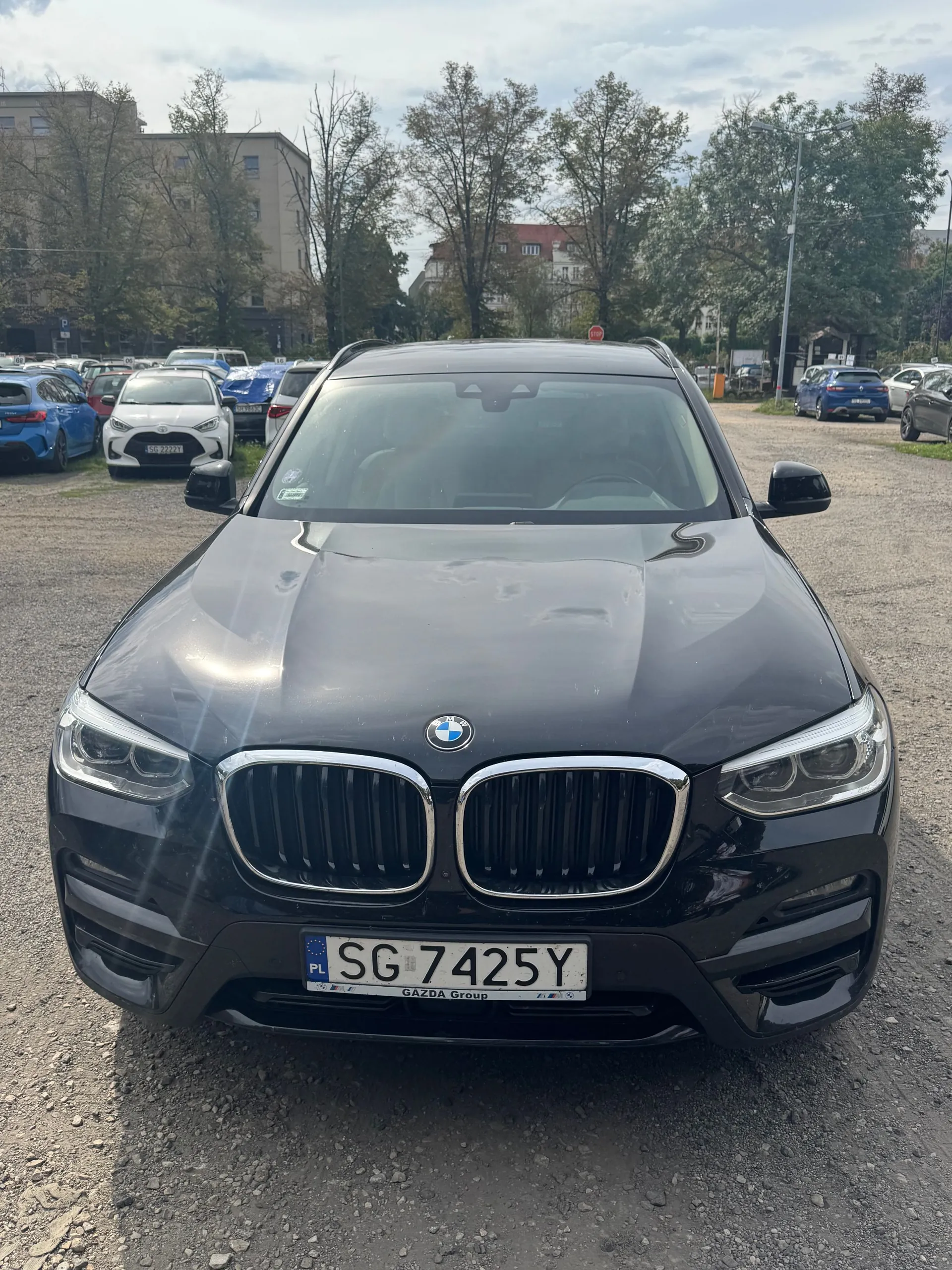 Czarny samochód marki BMW zaparkowany przodem do obiektywu na żwirowym parkingu. Widoczna jest charakterystyczna atrapa chłodnicy w kształcie nerek oraz tablica rejestracyjna o numerze SG 7425Y z oznaczeniem PL. W tle znajdują się inne zaparkowane samochody, drzewa oraz budynki mieszkalne pod zachmurzonym niebem.