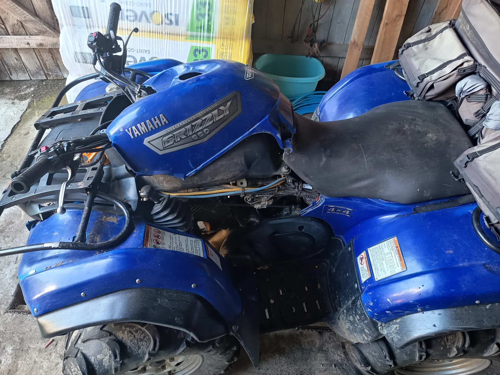 Niebieski quad Yamaha Grizzly 600 zaparkowany w garażu. Na zbiorniku paliwa widoczne napisy 'YAMAHA' oraz 'GRIZZLY 600'. Czarne siedzenie, kierownica, opony z brudem i naklejki ostrzegawcze na błotnikach. W tle drewniana ściana, niebieski pojemnik, opakowanie z napisem 'ISOVER' oraz cyfrą '35'.