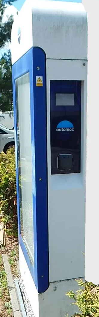 Biały automat parkingowy z niebieskimi elementami wykończeniowymi. Na przednim panelu znajduje się mały ekran, logo z napisem 'automoc' oraz otwór na bilet lub kartę. Po lewej stronie widoczna jest szklana witryna z informacjami. Urządzenie stoi na zewnątrz, w tle widać fragment białego samochodu oraz zielone krzewy.