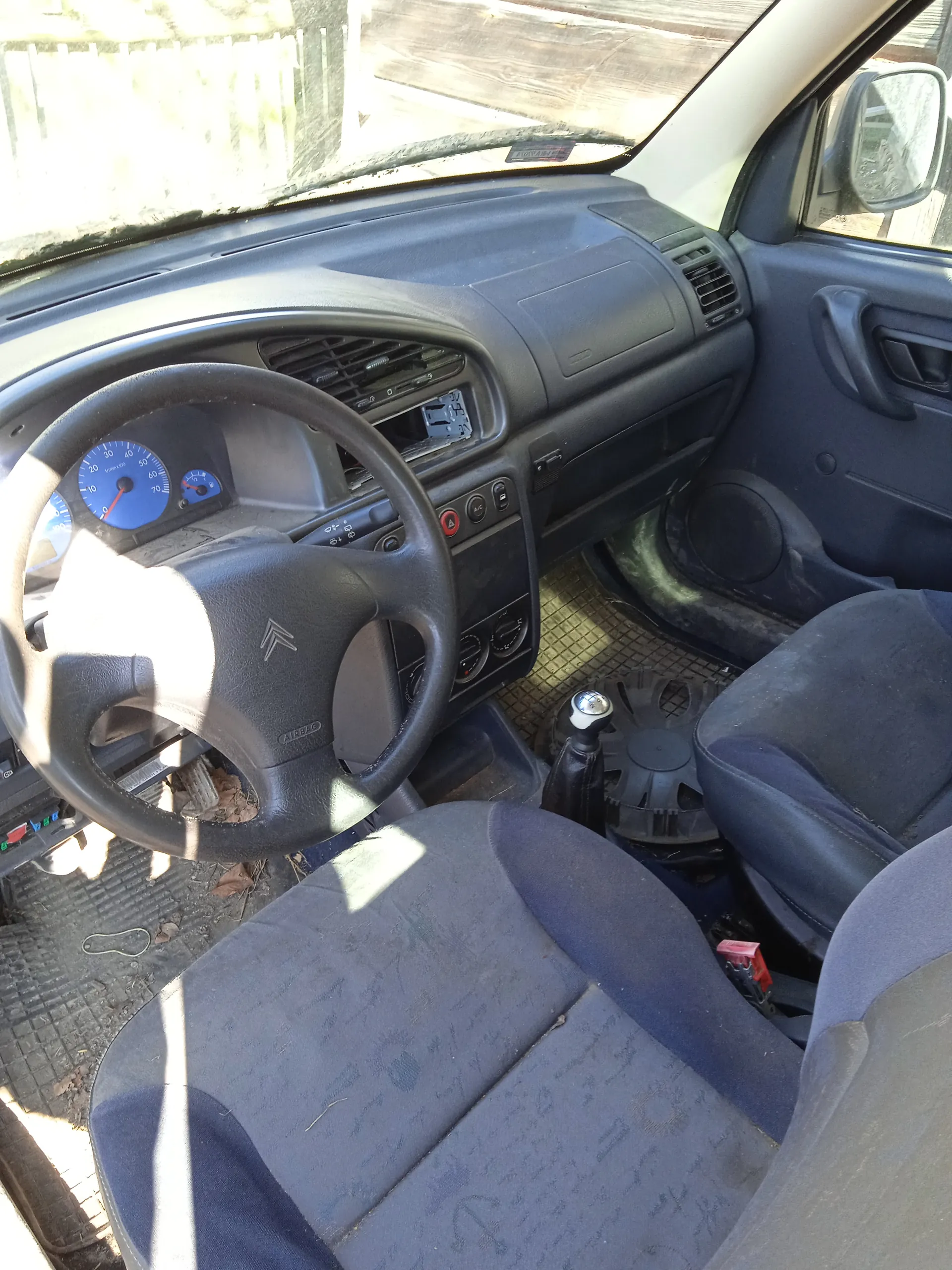 Wnętrze samochodu z widocznym kierownicą marki Citroën, niebieskimi wskaźnikami prędkościomierza i obrotomierza na desce rozdzielczej, ciemnoszarym panelem deski rozdzielczej, dźwignią zmiany biegów, ciemnoniebieskimi siedzeniami z widocznymi śladami zużycia oraz brudnymi dywanikami podłogowymi. Przez przednią szybę widoczny jest fragment ogrodzenia i drewnianego obiektu na zewnątrz. Wnętrze oświetlone jest światłem słonecznym.