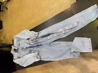 Jasnoniebieskie dżinsowe spodnie z kombinezonem leżące na ciemnej powierzchni. Spodnie mają przyciski na przodzie, kieszenie oraz rozłożone nogawki. Tło składa się z jasnej podłogi i części ciemnego mebla.