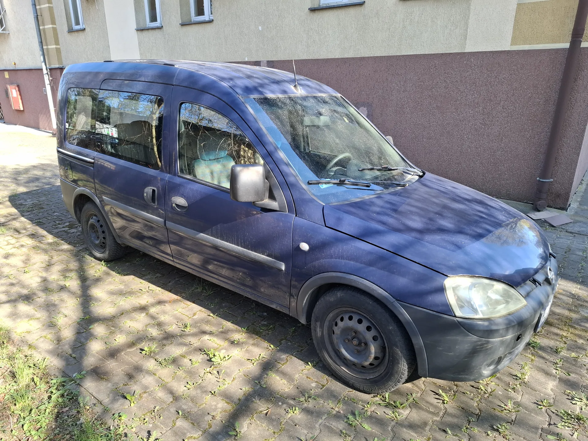 Ciemnoniebieski furgonetka zaparkowana na brukowanej powierzchni z widocznymi szczelinami porośniętymi trawą. Pojazd ma czarne felgi, brudną karoserię i przednią szybę z wycieraczkami. W tle budynki z beżowymi i brązowymi ścianami oraz oknami. Cień drzewa pada na samochód i bruk.