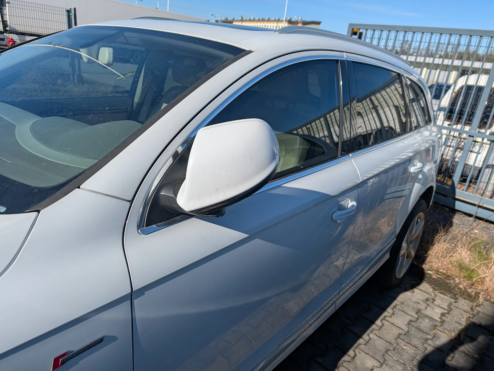 Srebrny samochód typu SUV zaparkowany na brukowanej powierzchni. Widoczne są boczne lusterko, okna, drzwi z klamką oraz czerwony emblemat na przednim błotniku. W tle metalowe ogrodzenie, budynek i jasne niebo.