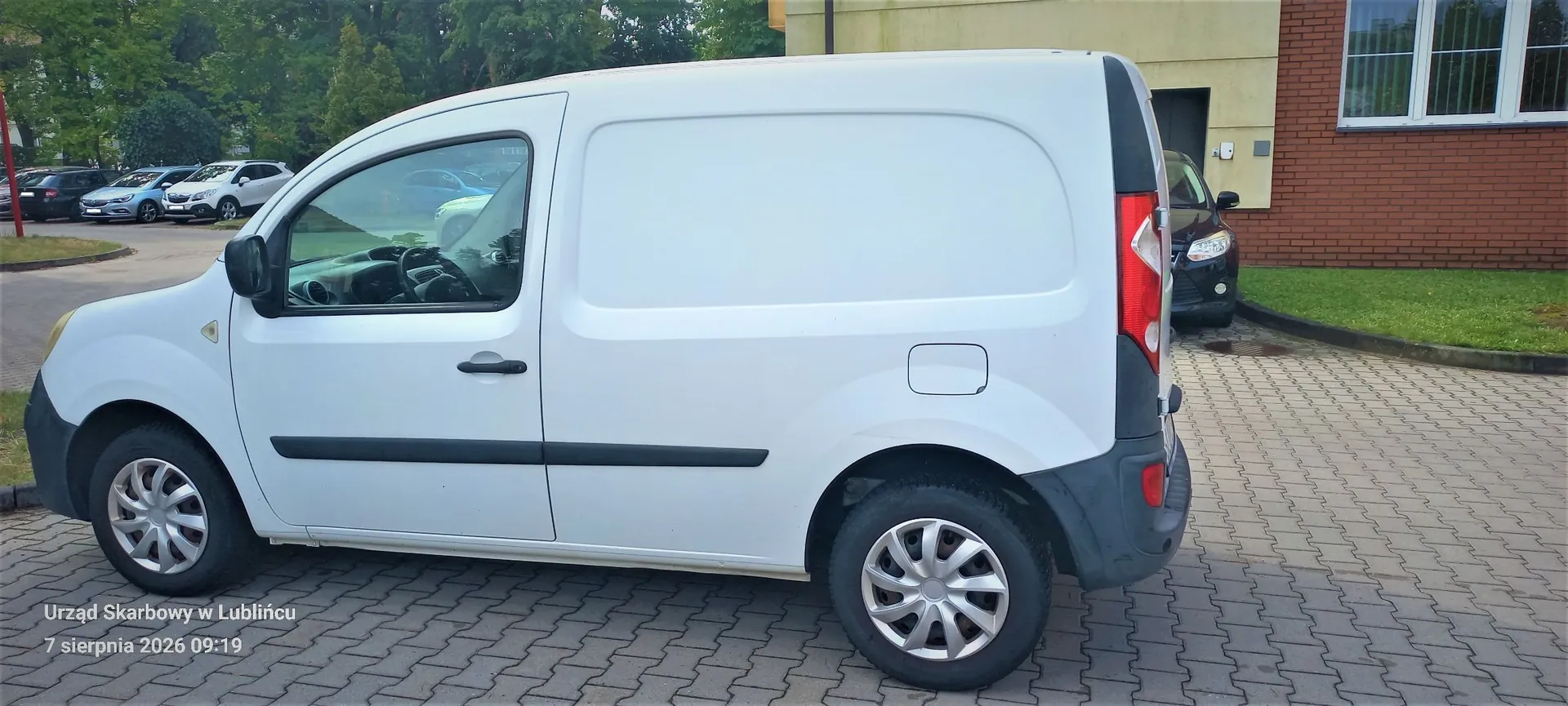 Biały samochód dostawczy zaparkowany na kostce brukowej przed budynkiem z czerwonej cegły. Pojazd jest widoczny z profilu od strony kierowcy. W tle widać inne zaparkowane samochody oraz zieleń. W lewym dolnym rogu obrazu znajduje się biały napis: 'Urząd Skarbowy w Lublińcu 7 sierpnia 2026 09:19'.