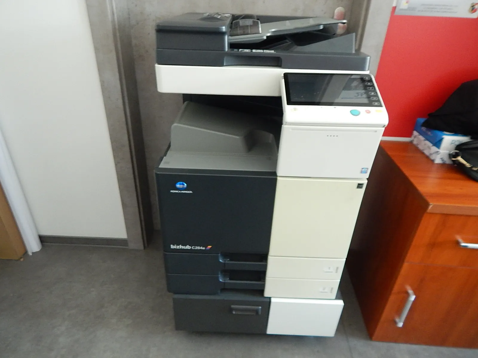 Wielofunkcyjna drukarka biurowa marki Konica Minolta, model bizhub C284e, w kolorach białym, szarym i czarnym. Urządzenie stoi w pomieszczeniu obok drewnianej szafki. Na górze znajduje się skaner z automatycznym podajnikiem dokumentów oraz panel sterowania z ekranem dotykowym.