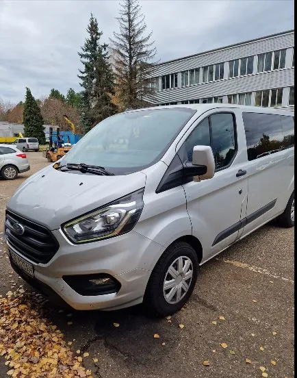 Srebrny samochód dostawczy marki Ford Transit Custom zaparkowany na asfaltowym placu. W tle widoczny jest nowoczesny, biały budynek biurowy z wieloma oknami, wysokie drzewa iglaste oraz żółty wózek widłowy. Na ziemi leżą jesienne, żółte liście.
